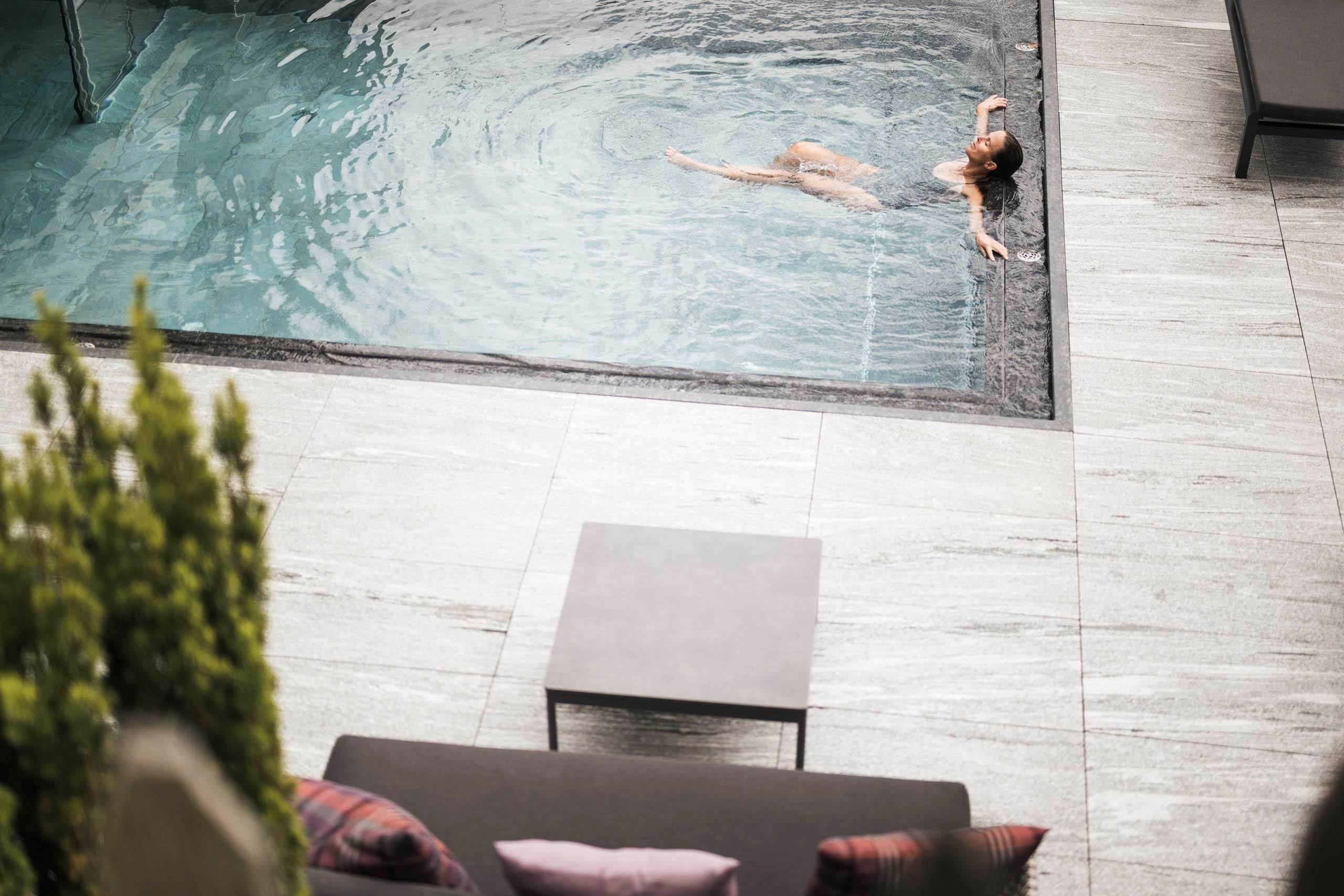 An elegant outdoor area with loungers and umbrellas, surrounded by trees and mountains. The pool reflects the sun and provides a relaxing atmosphere.