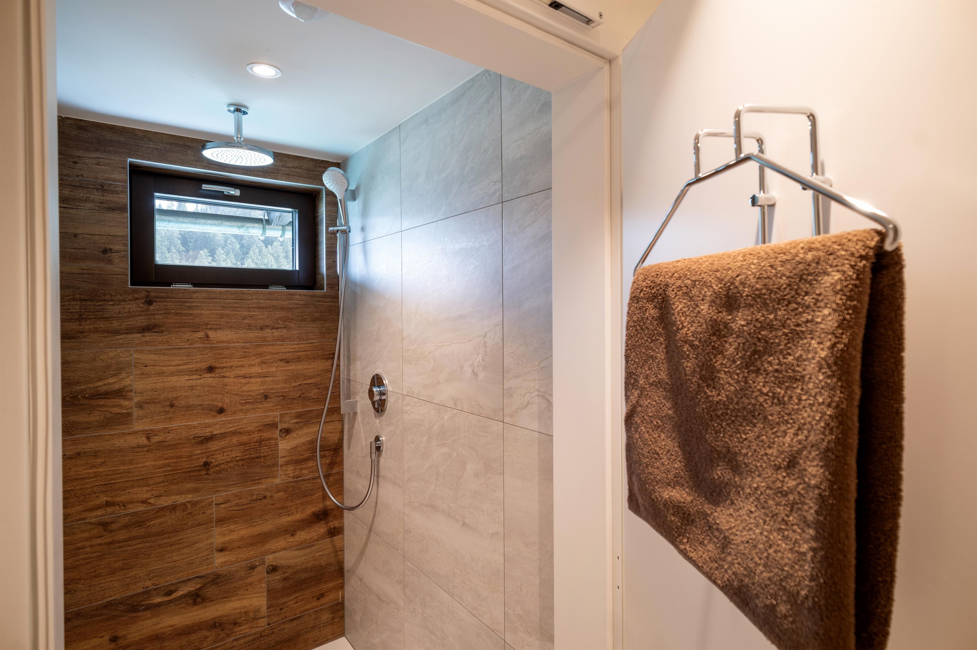 A modern shower with tiles and a wooden cladding. A towel hangs on a hook in the corner.