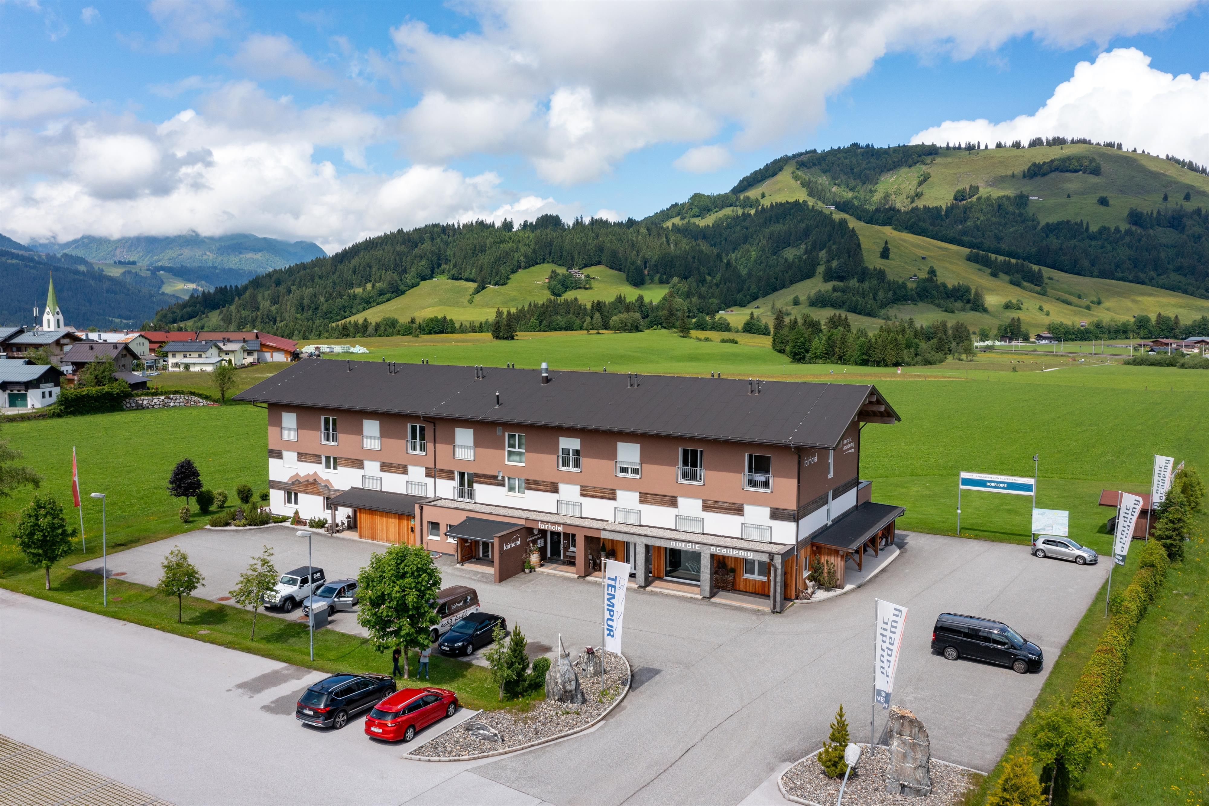 A modern building in a rural setting with green meadows and mountains in the background. Several cars are parked in the parking lot.