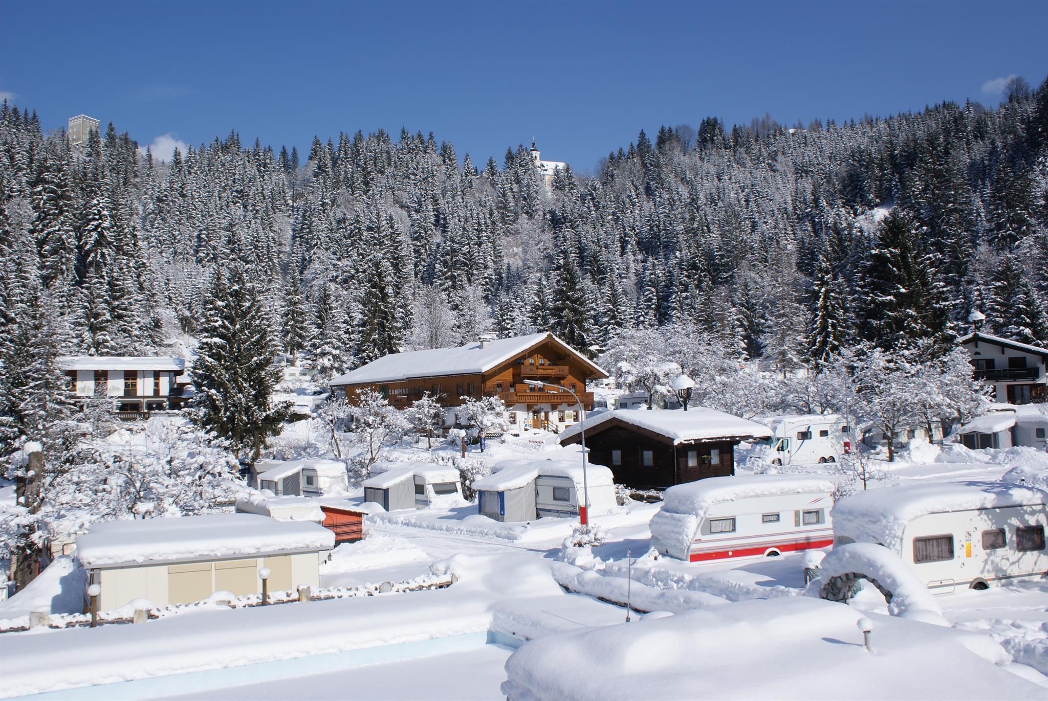 A snowy landscape with wooden houses and caravans. Tall, snow-covered trees surround the scene under a clear blue sky.