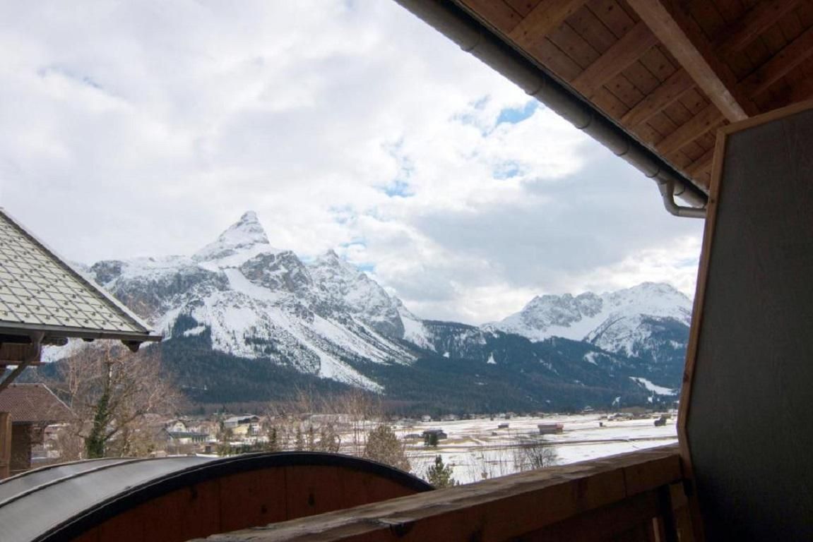 A view of snow-covered mountains under a cloudy sky. In the landscape, there are some buildings and trees visible.