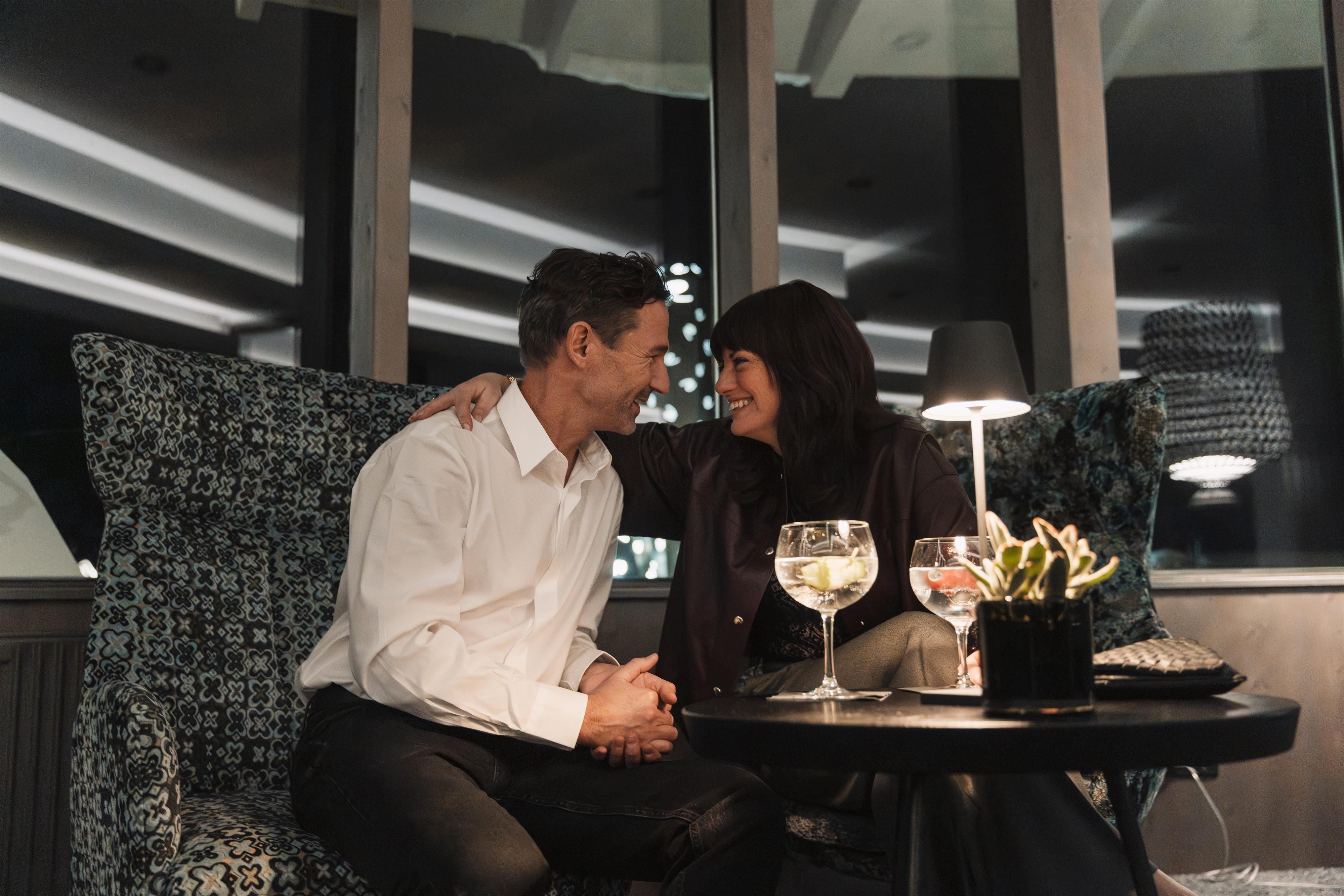 A romantic couple sits in a cozy lounge. They smile at each other and enjoy drinks in the dim light.