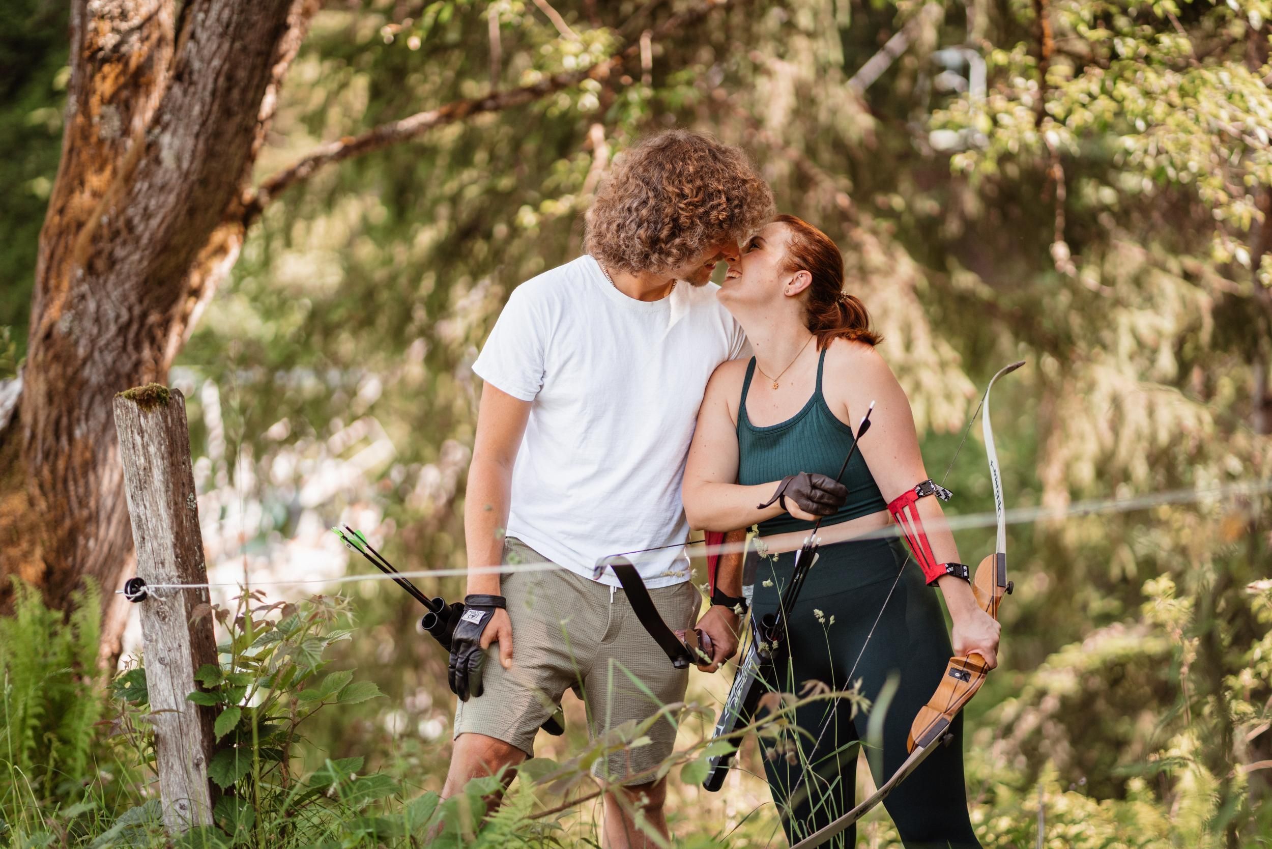 A young couple stands in nature, surrounded by trees. They are smiling at each other and seem to share a romantic connection.