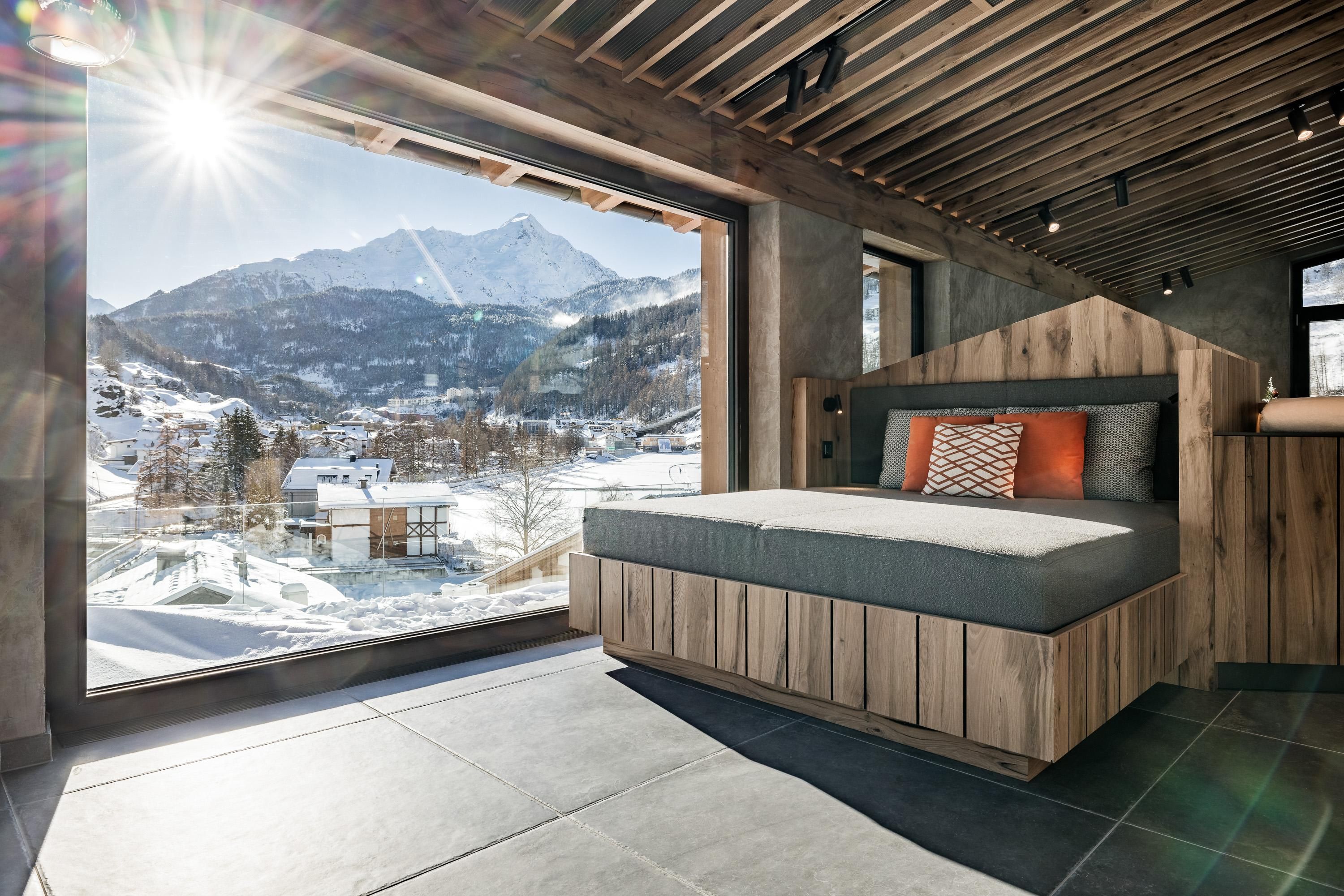 A modern bedroom with a large window and a view of snow-capped mountains. The sun is shining, and there is a cozy chaise lounge with colorful pillows.