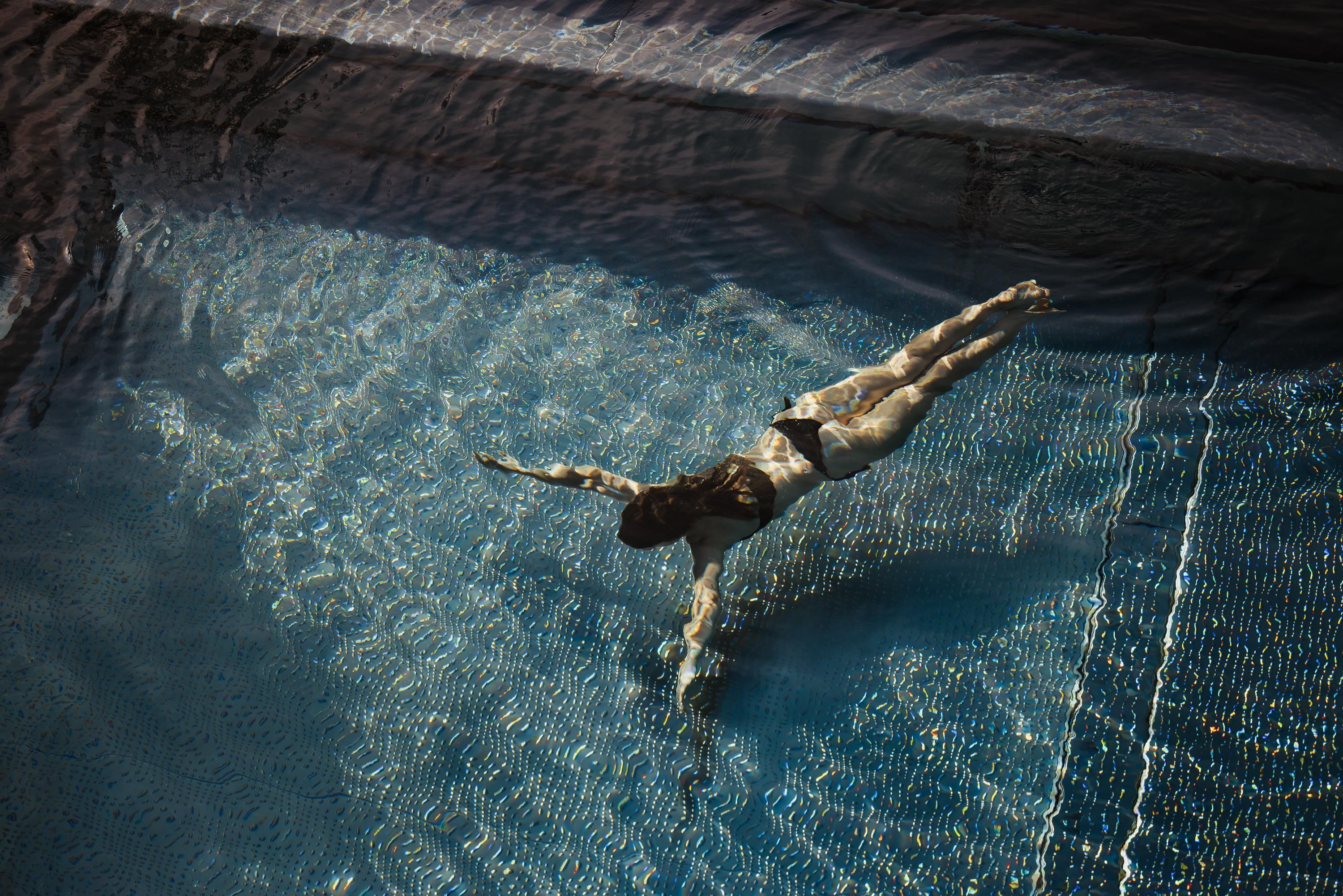 A person is diving in a clear, blue pool. The sunlight creates a beautiful reflection on the surface of the water.