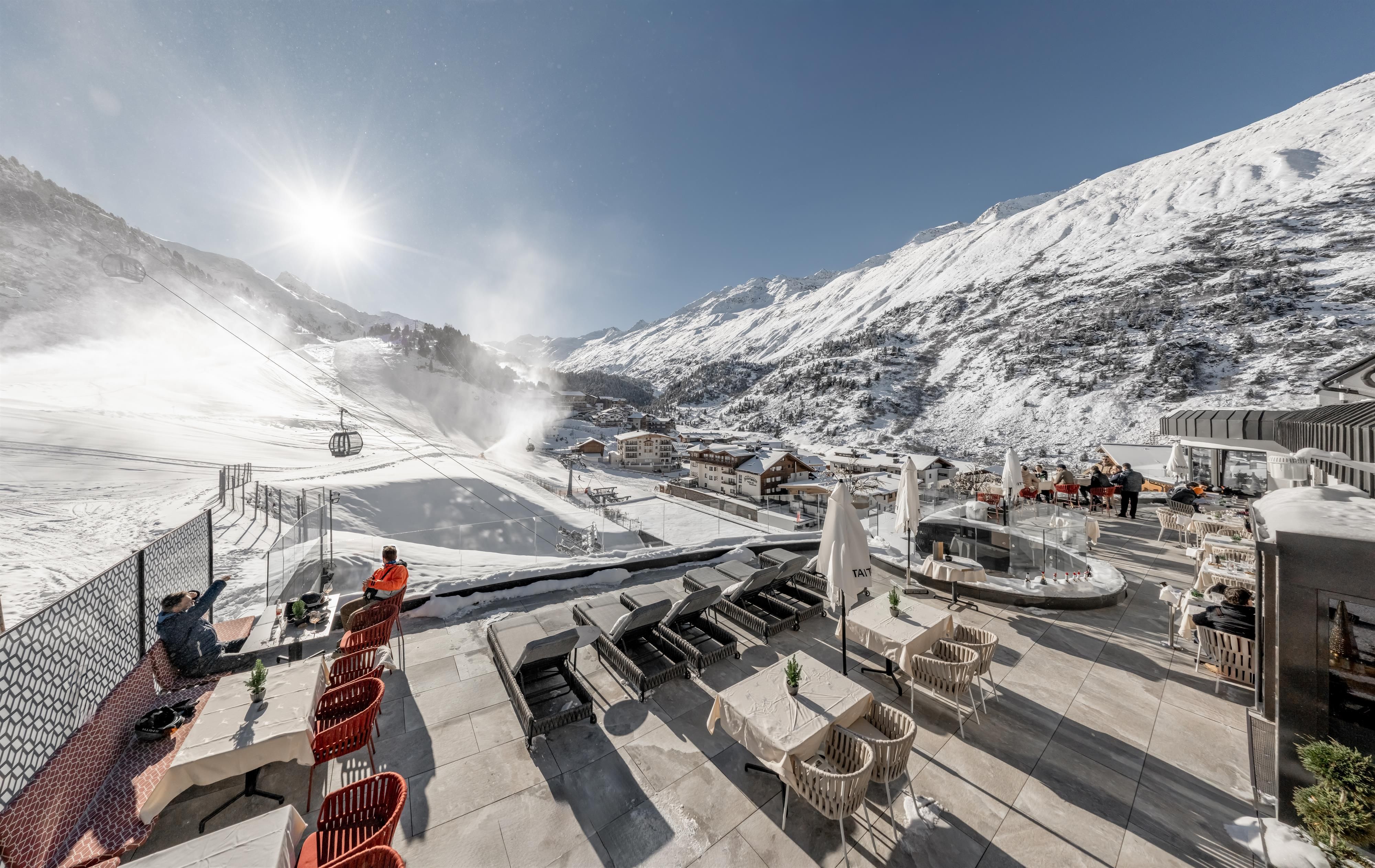 A mountain landscape full of snow with a clear blue sky. In the foreground, there are lounges and seating on a terrace.