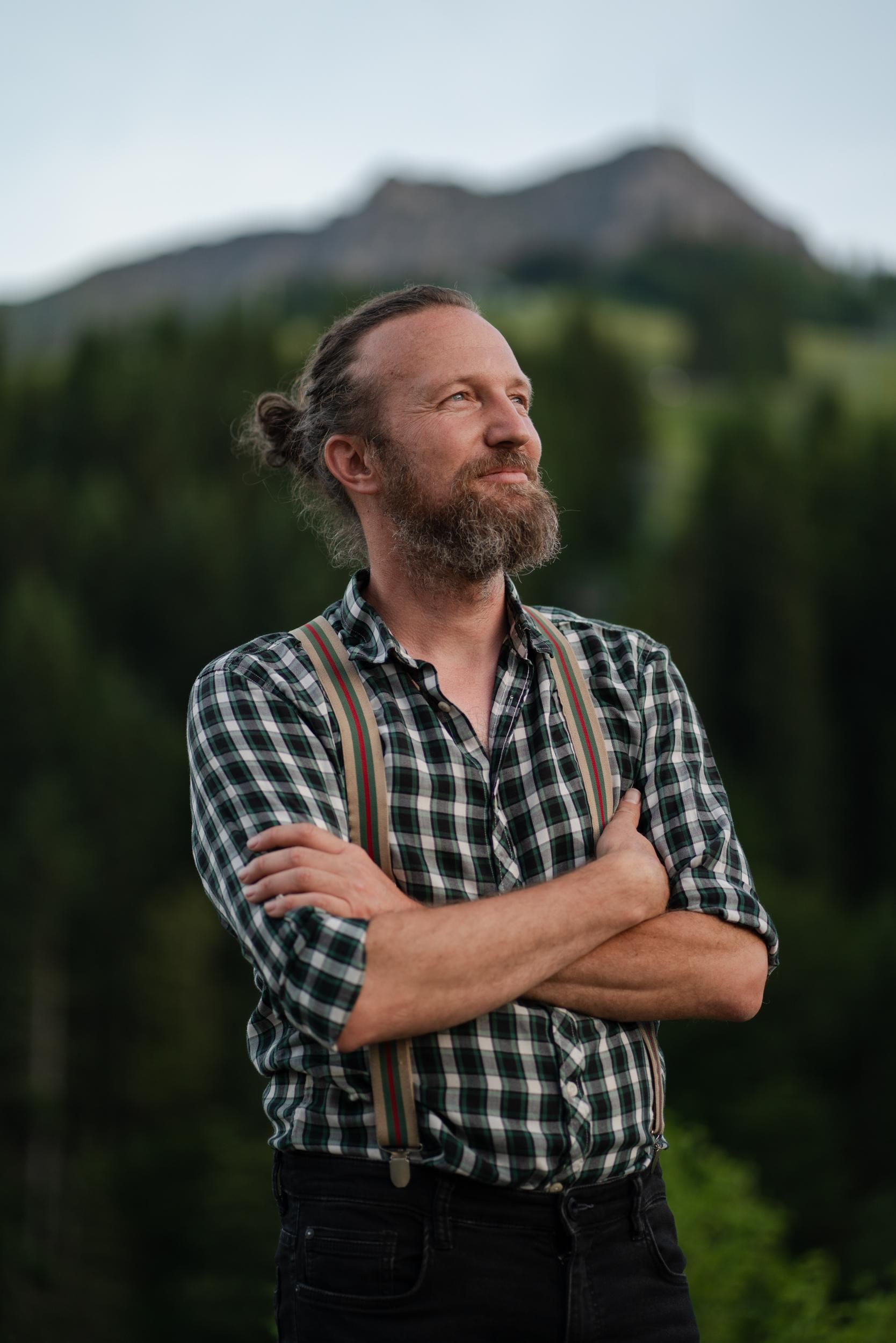 A man with a beard stands in nature and looks thoughtfully into the distance. In the background, trees and mountains can be seen.
