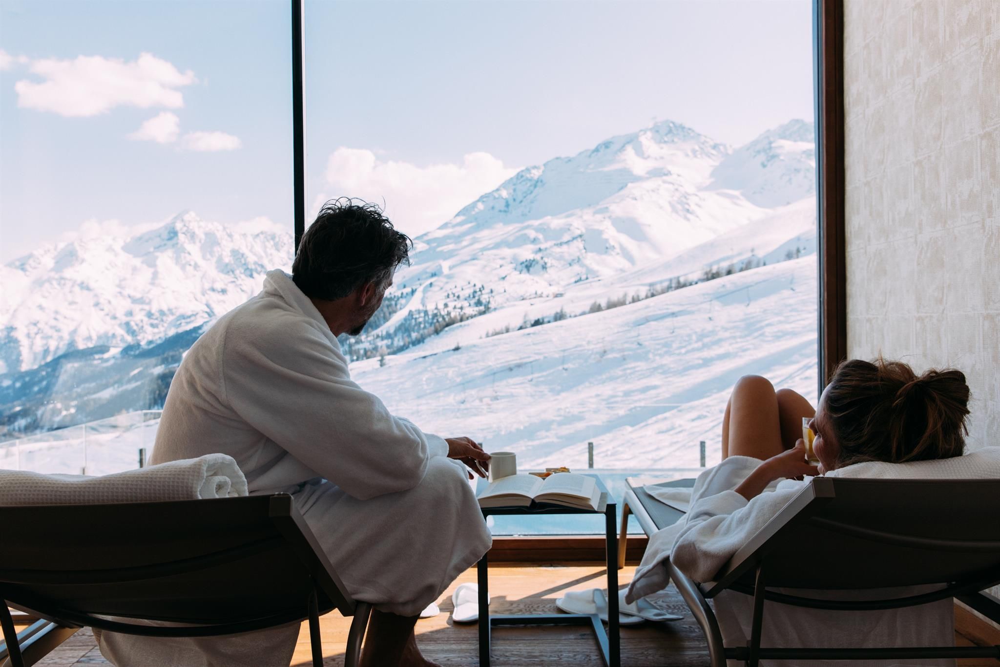 A couple in bathrobes relaxes in a modern room and enjoys the view of snow-covered mountains. The atmosphere radiates calm and coziness.
