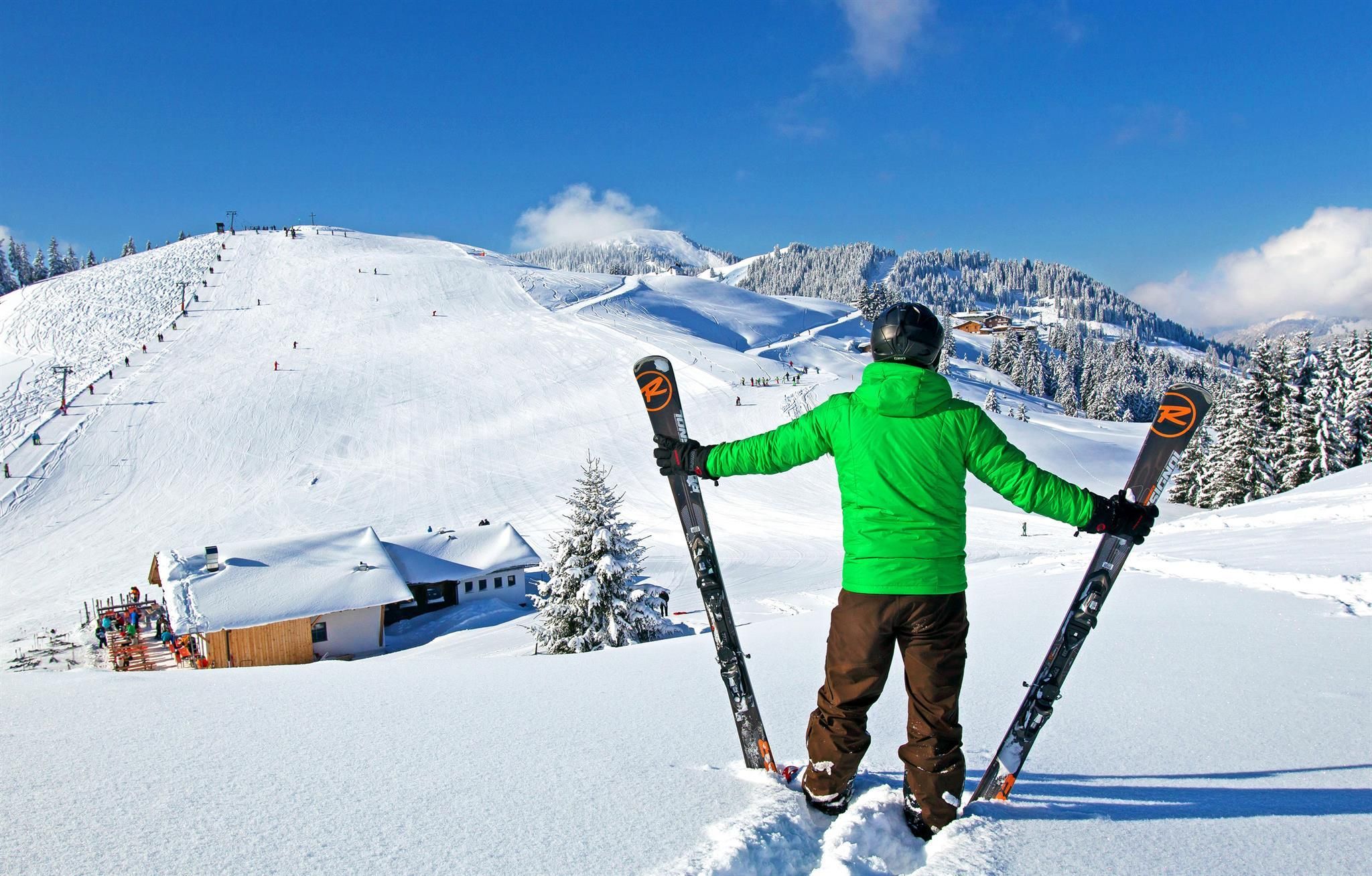 A skier stands on a snow-covered slope and looks out over the snowy landscape. In the background, there are lifts and a wooden house visible.