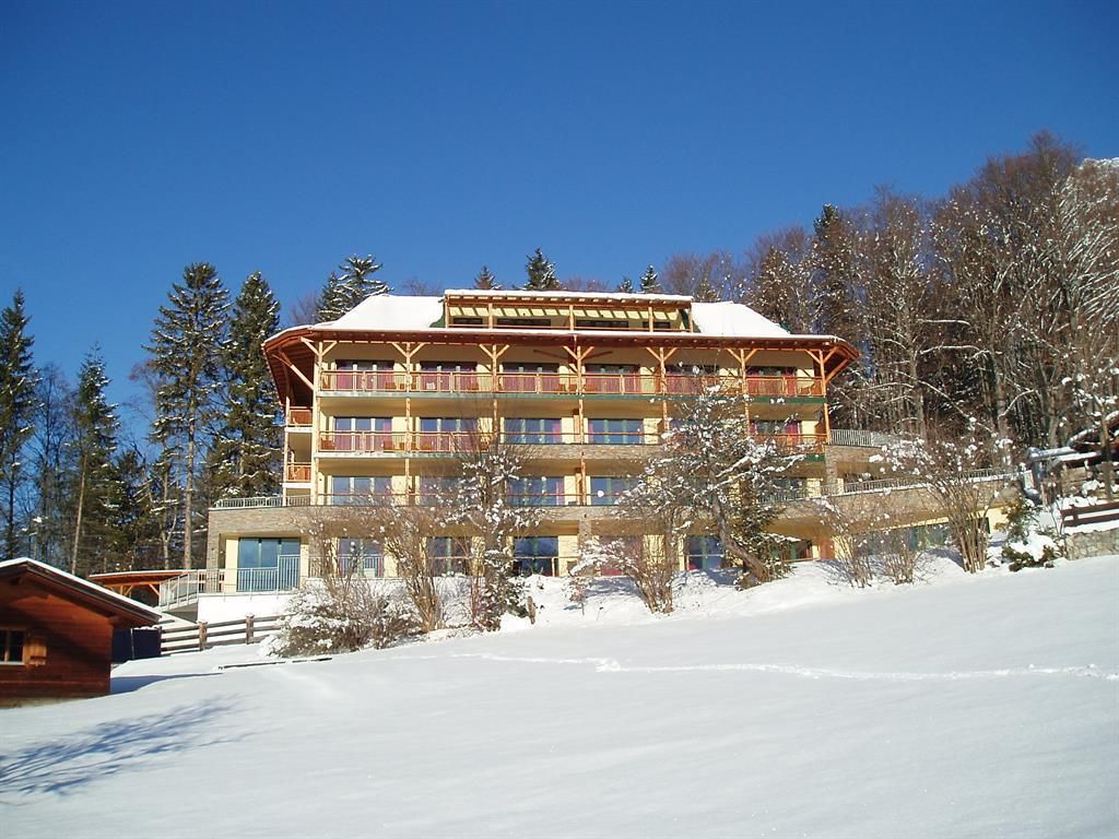 A cozy hotel in winter with snow-covered grounds. Surrounded by trees and a clear sky.