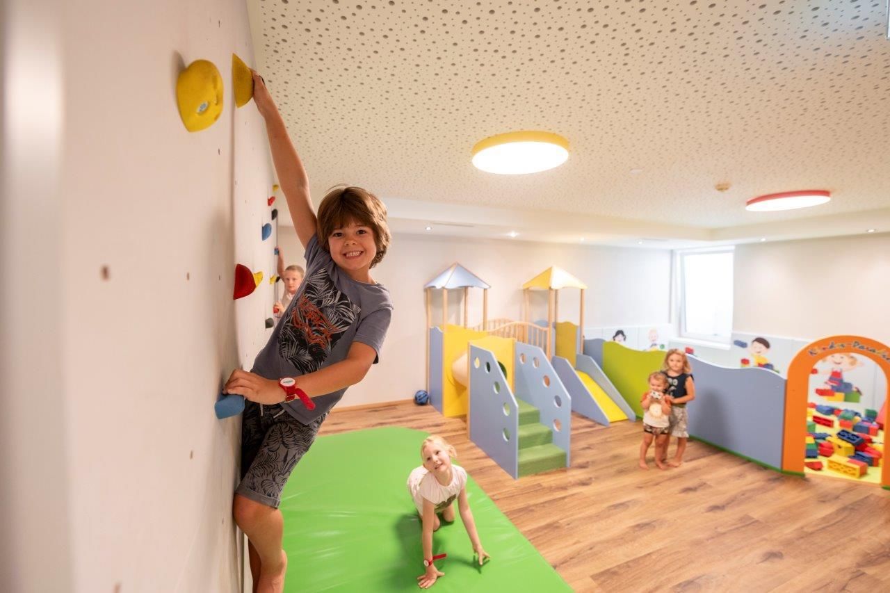 A lively play area with a climbing wall and play equipment for children. Several children are playing and having fun in a bright environment.