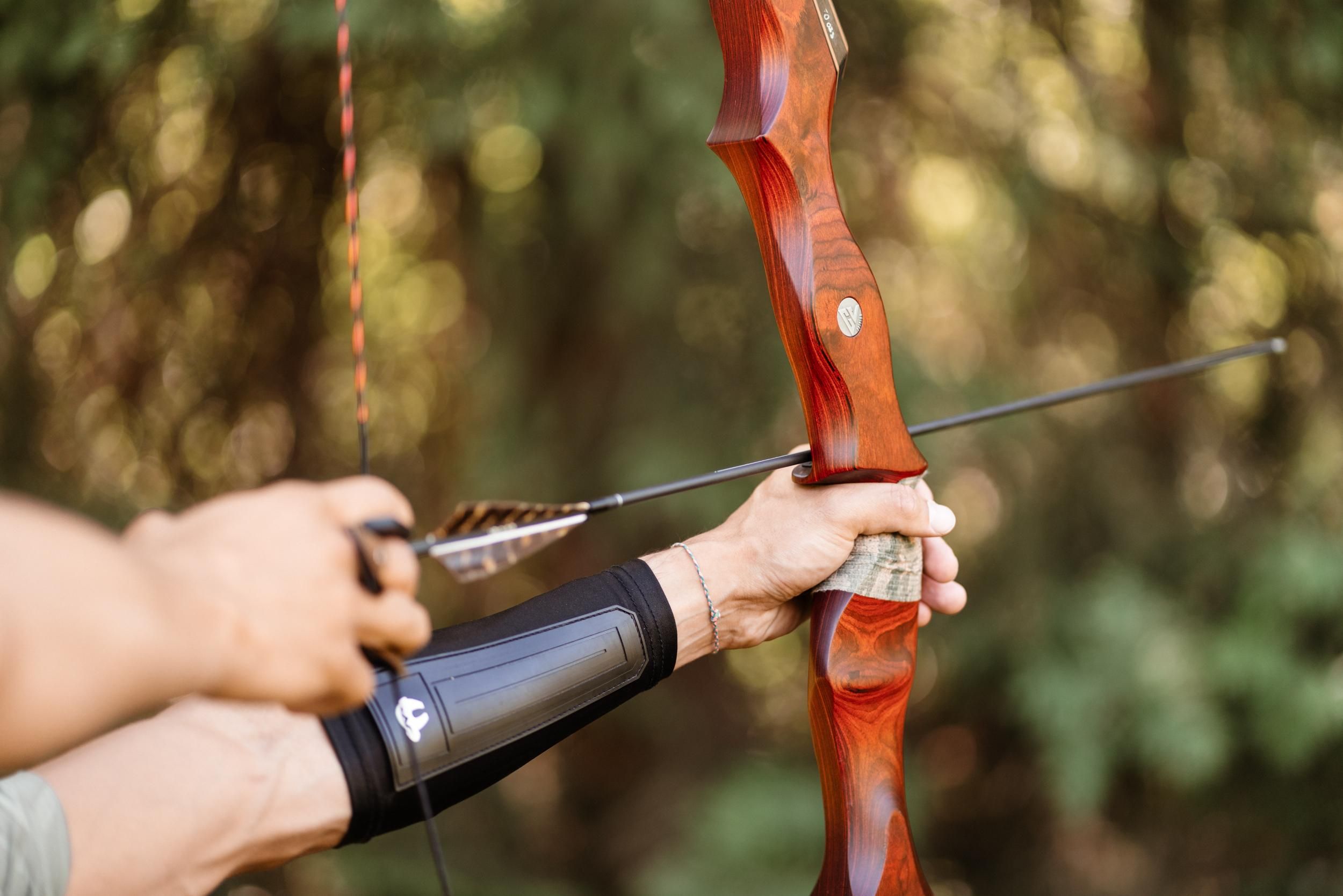 An archer aims with a traditional bow. The background shows green, blurred plants.