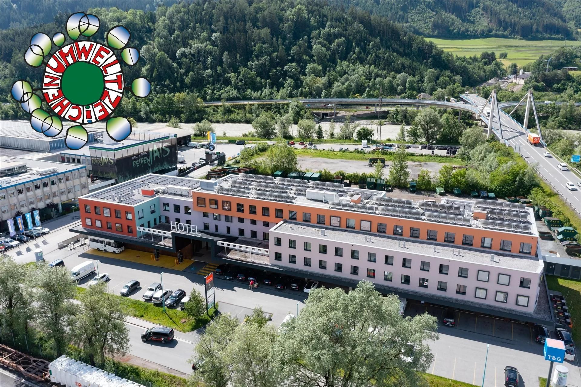 A modern building with various colors, surrounded by trees and a river. In the background, hills and a bridge can be seen.