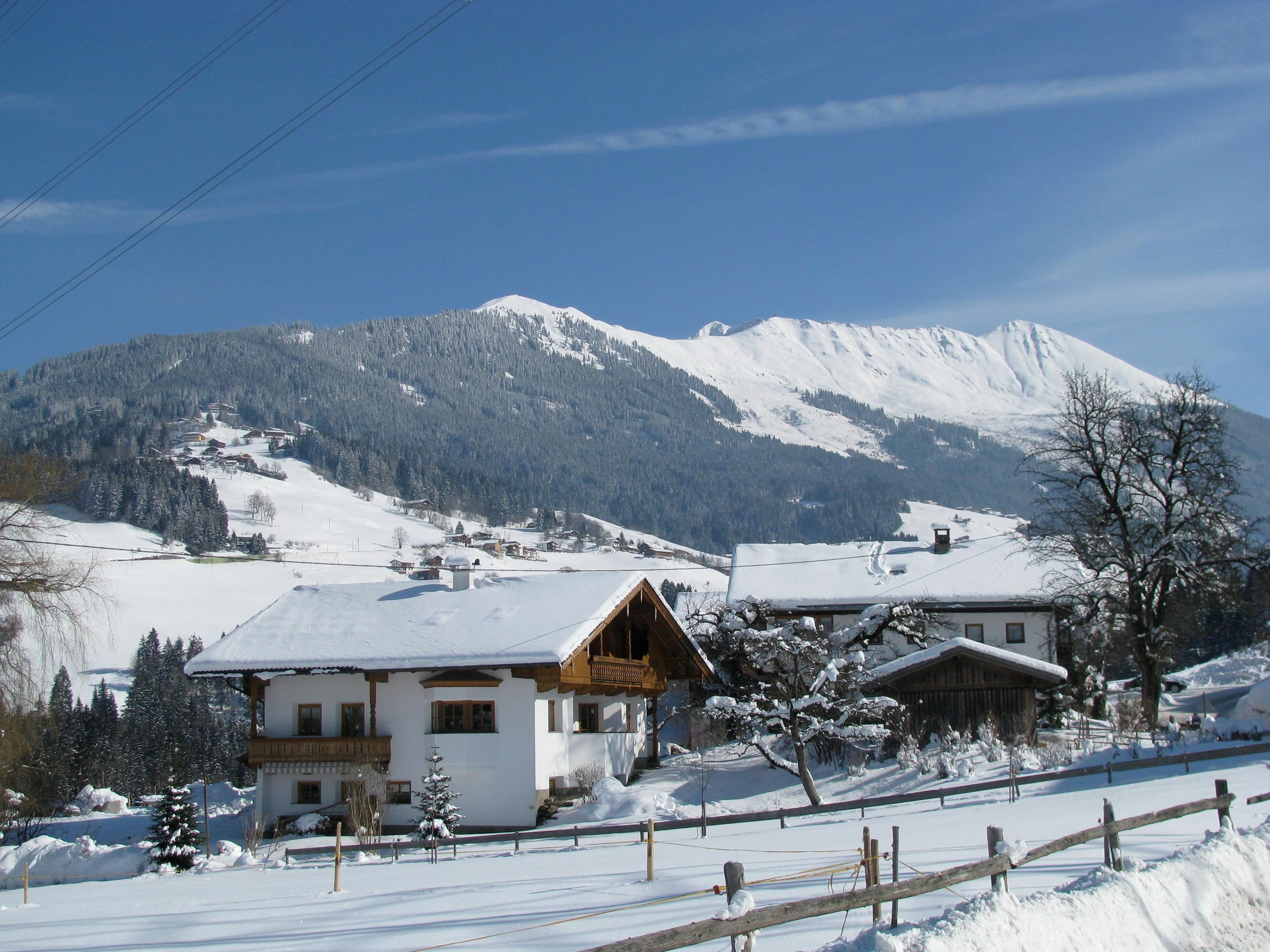 A snowy landscape with cozy houses and majestic mountains in the background. The clear sky and radiant sun give the scene a peaceful atmosphere.