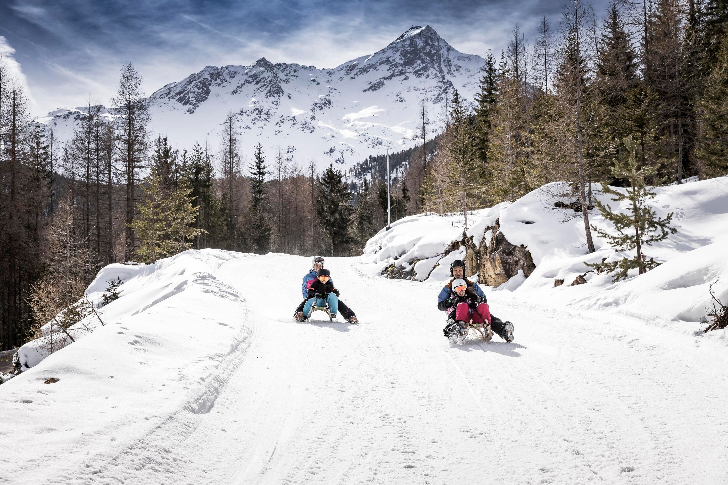 Two people are sledding on a snowy road in the mountains. In the background, there are tall mountains and a clear sky visible.