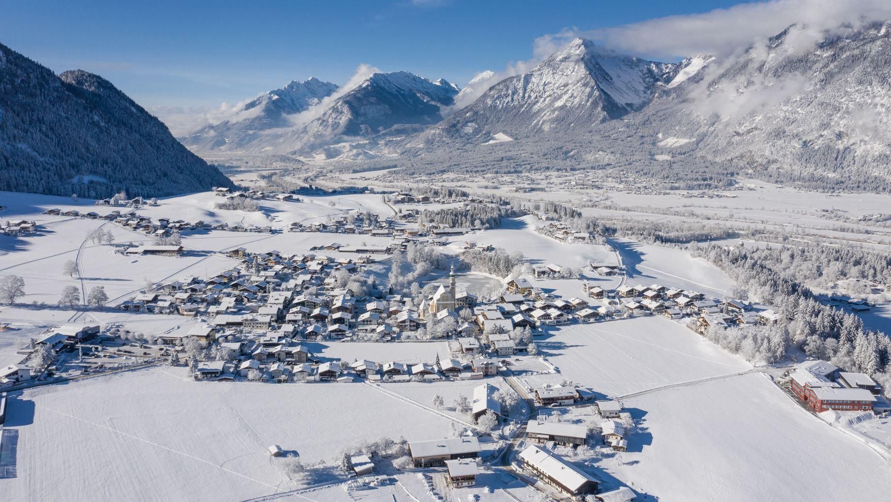 A picturesque winter landscape with snow-covered fields and houses. In the background, majestic mountains rise under a clear sky.