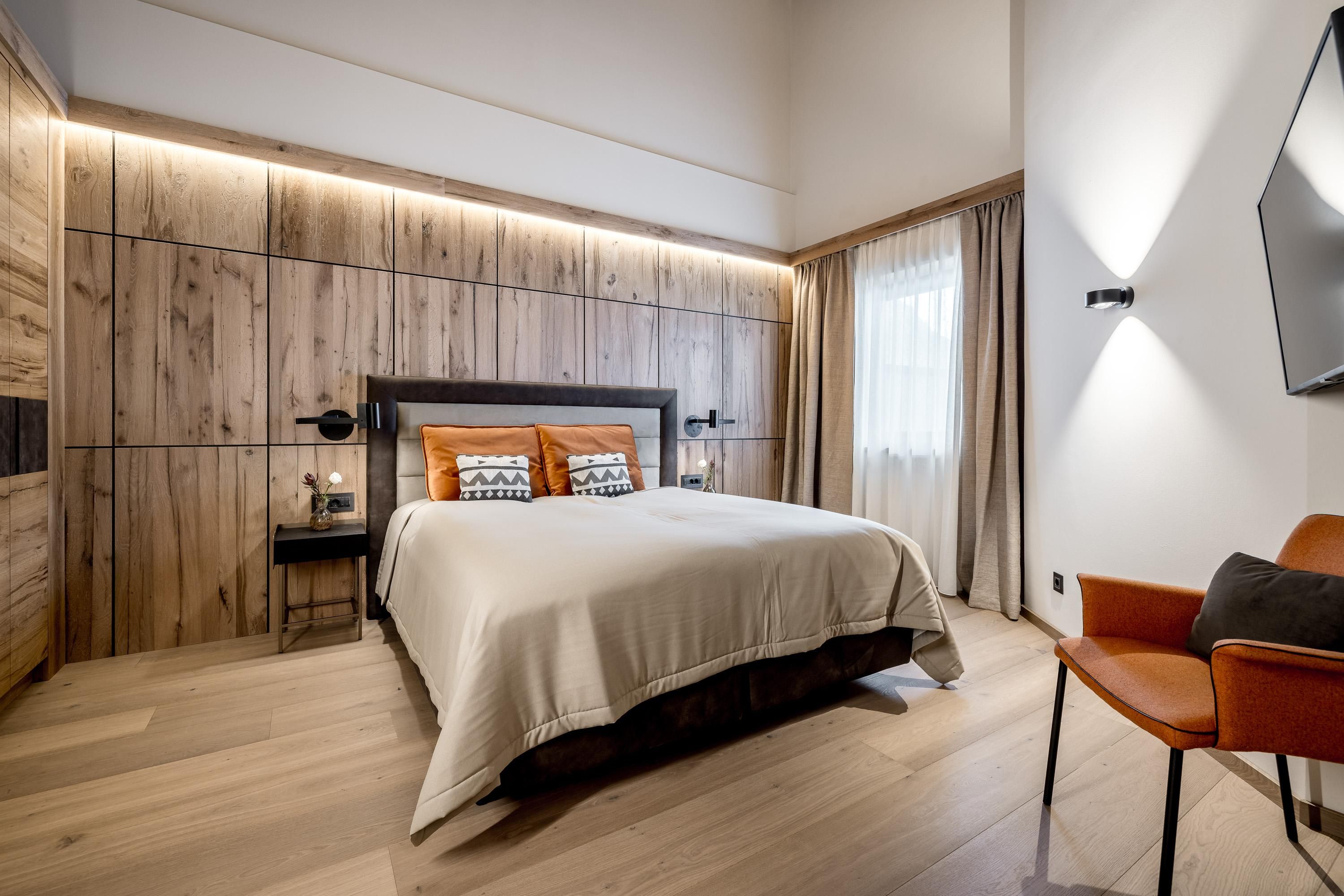 A modern bedroom with wooden paneling and soft lighting. A bed with orange cushions and a chair complement the elegant decor.
