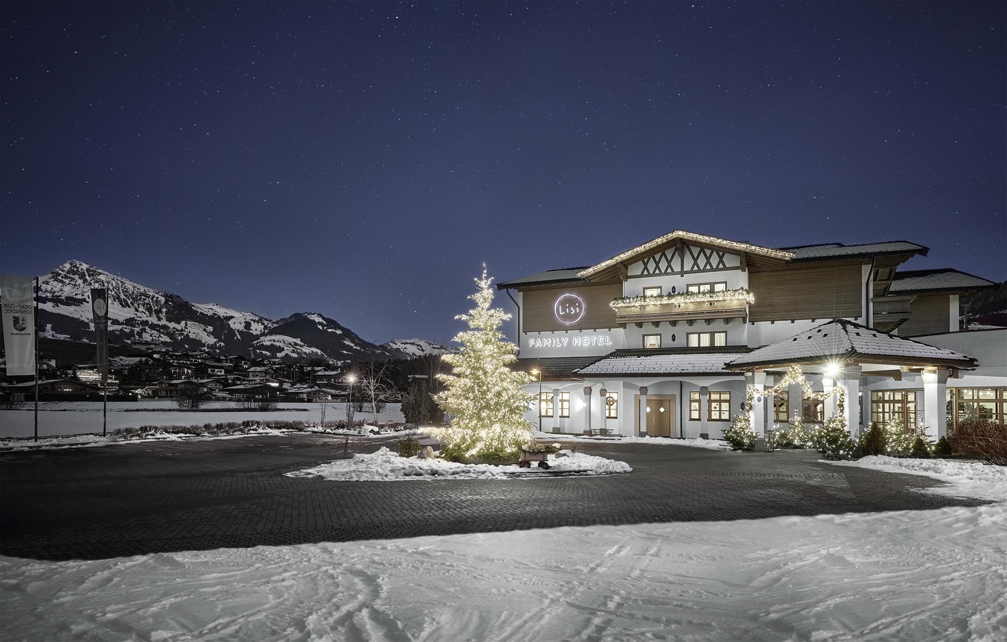 A hotel building in the snow at night, surrounded by mountains. A festively decorated Christmas tree is visible in the foreground.