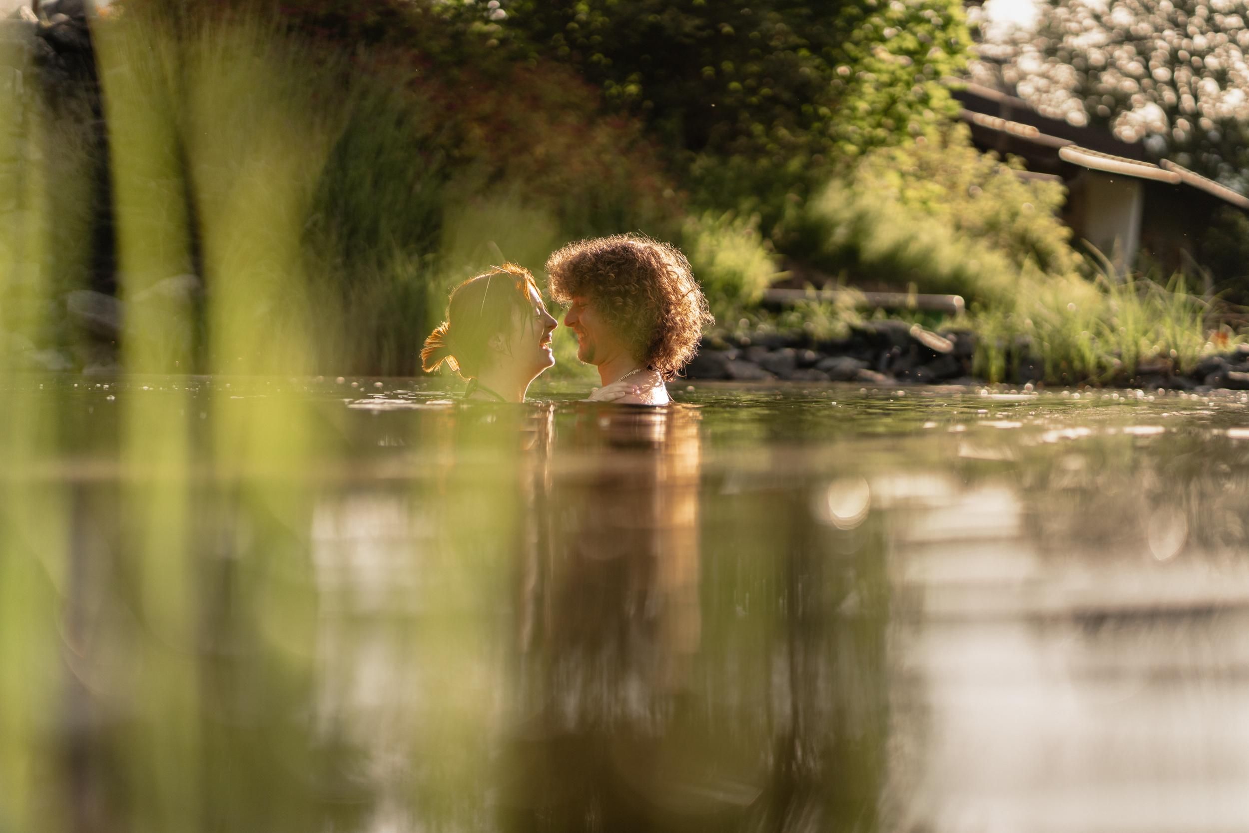 Two people are standing in the water and looking at each other. The surroundings are green and sunny, creating a relaxed atmosphere.
