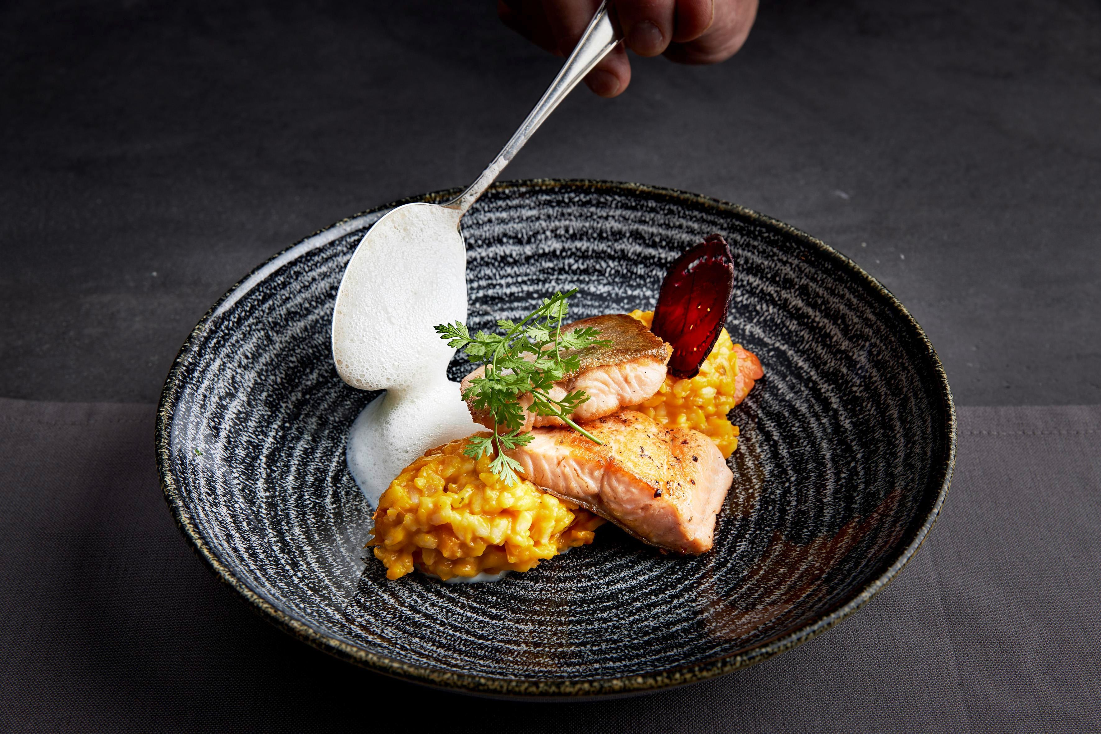 A delicious dish with salmon, served on a creamy risotto base. The dish is garnished with a white sauce and a sprig of herbs.