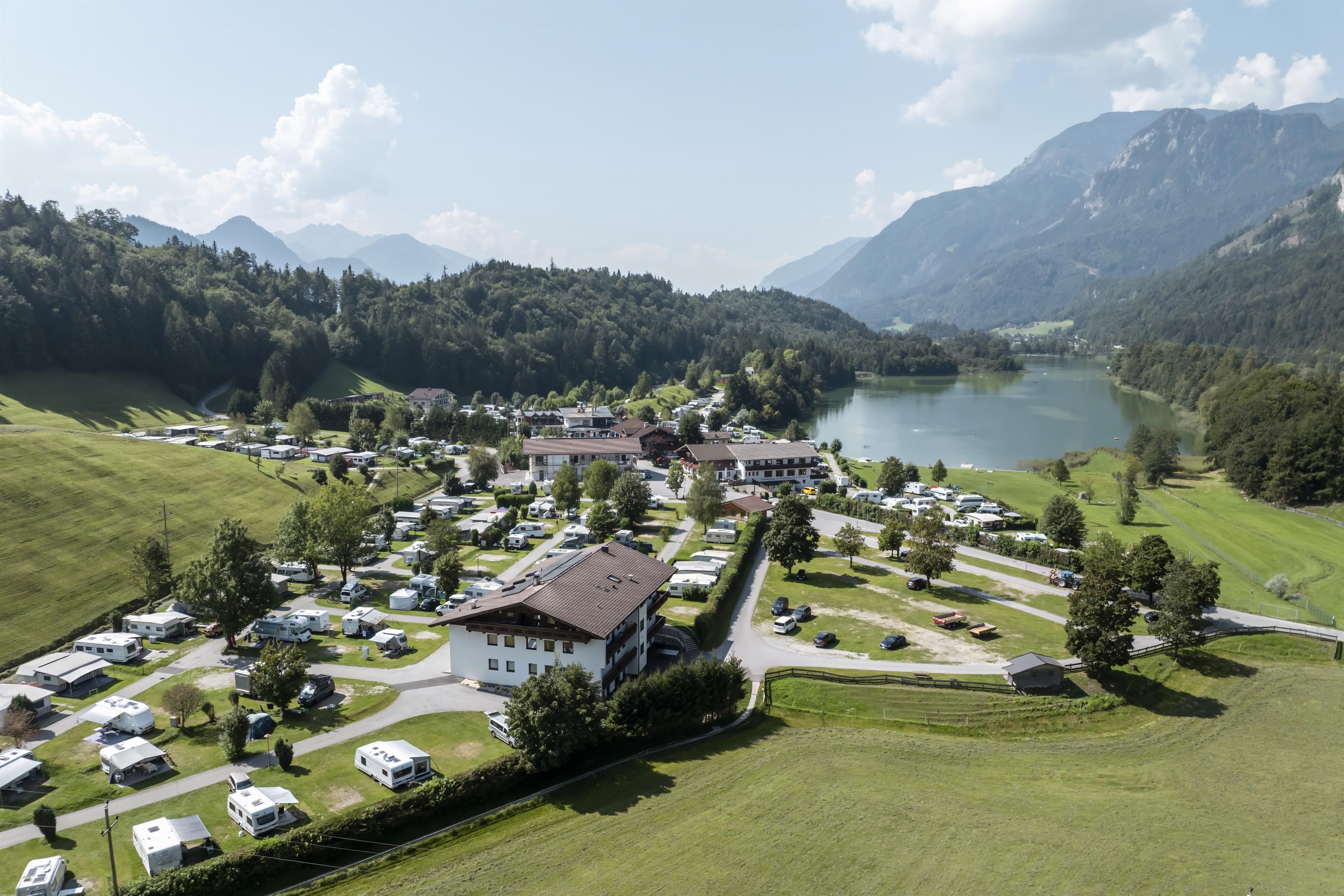 An idyllic landscape with a campsite, surrounded by green meadows and mountains. In the background, there is a clear lake under a blue sky.
