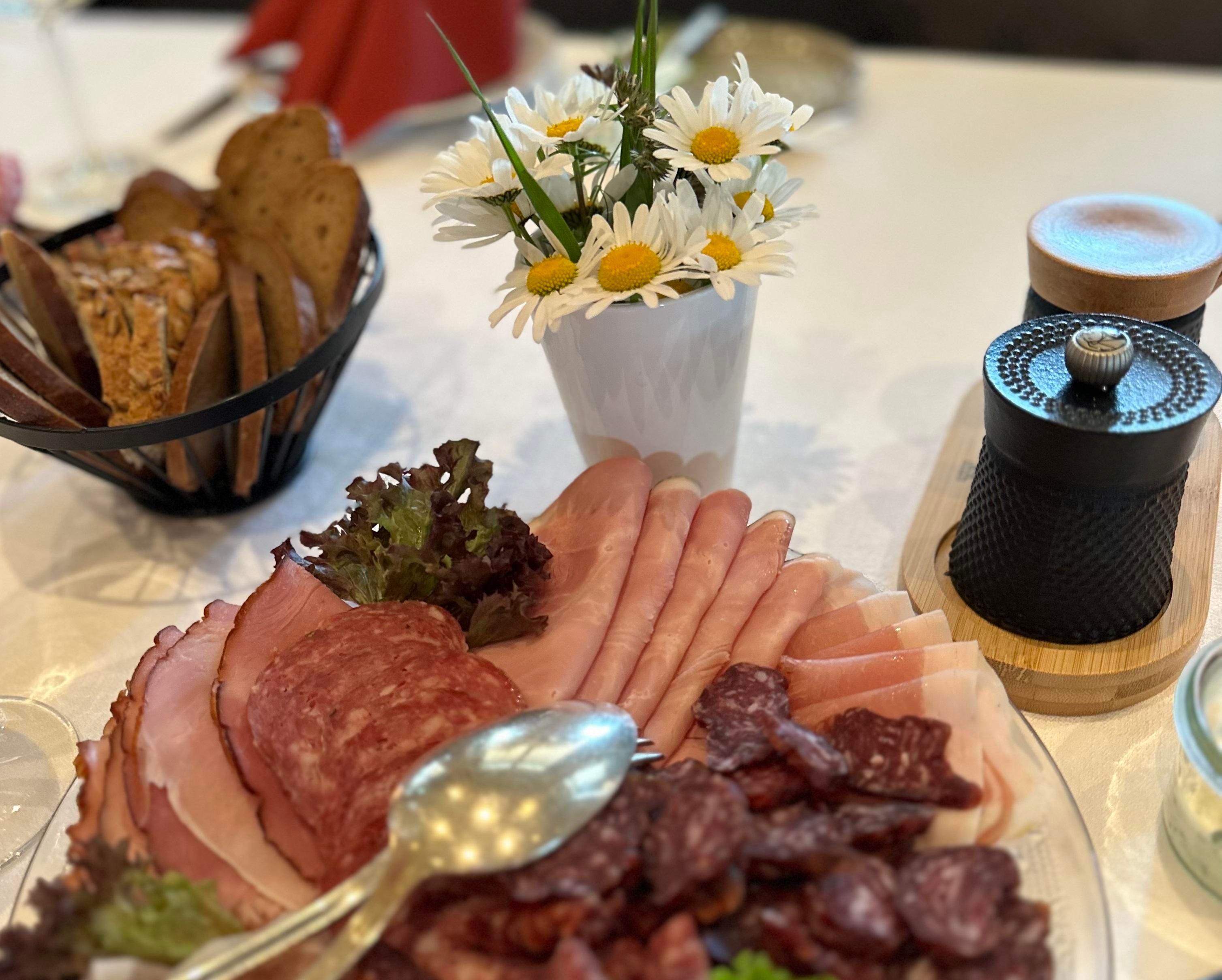 A beautiful plate with various cold cuts, decorated with salad. In the background is a small vase with daisies.