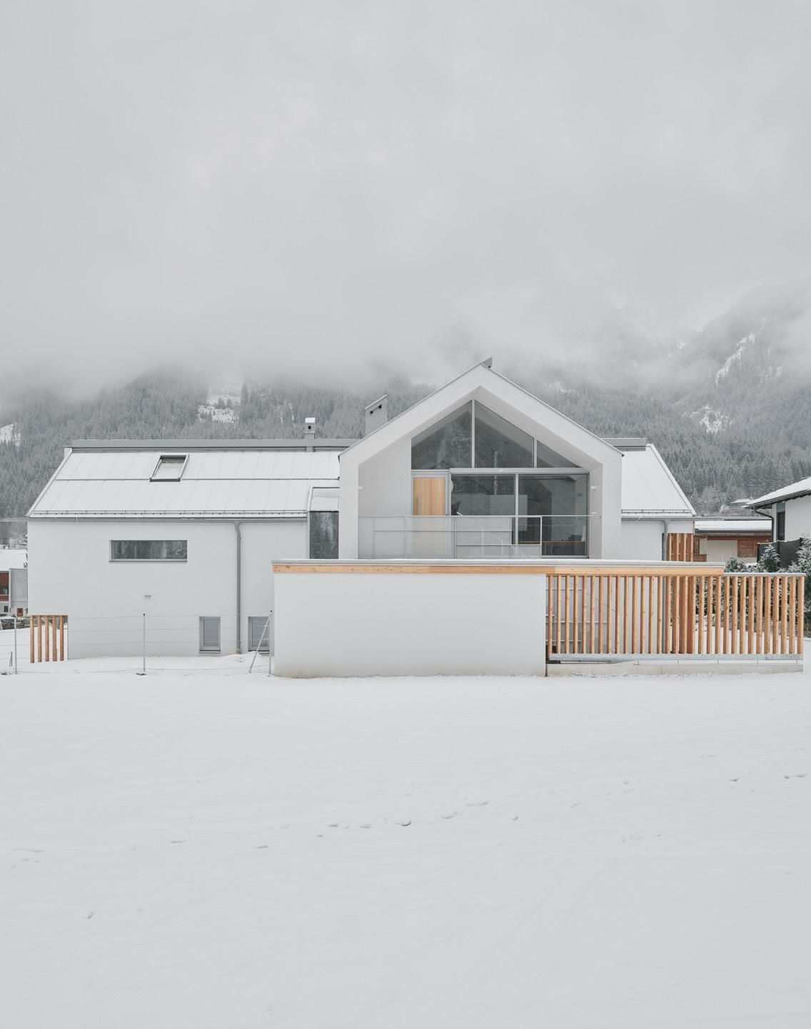 A modern house in the snow, surrounded by a winter landscape. The sky is overcast and the mountains are visible in the background.