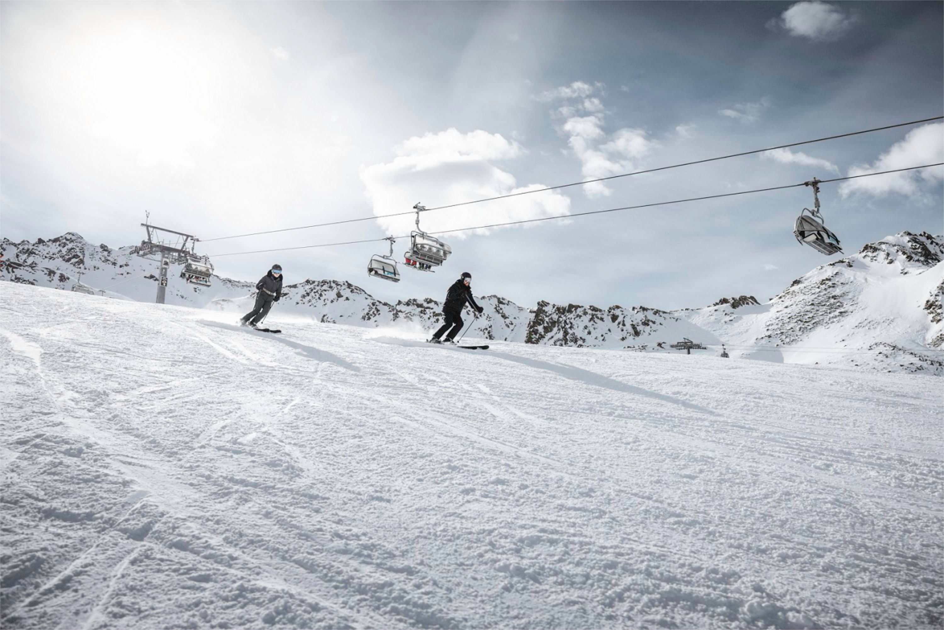 Two skiers are skiing down a snow-covered slope. In the background, lifts and mountains are visible.