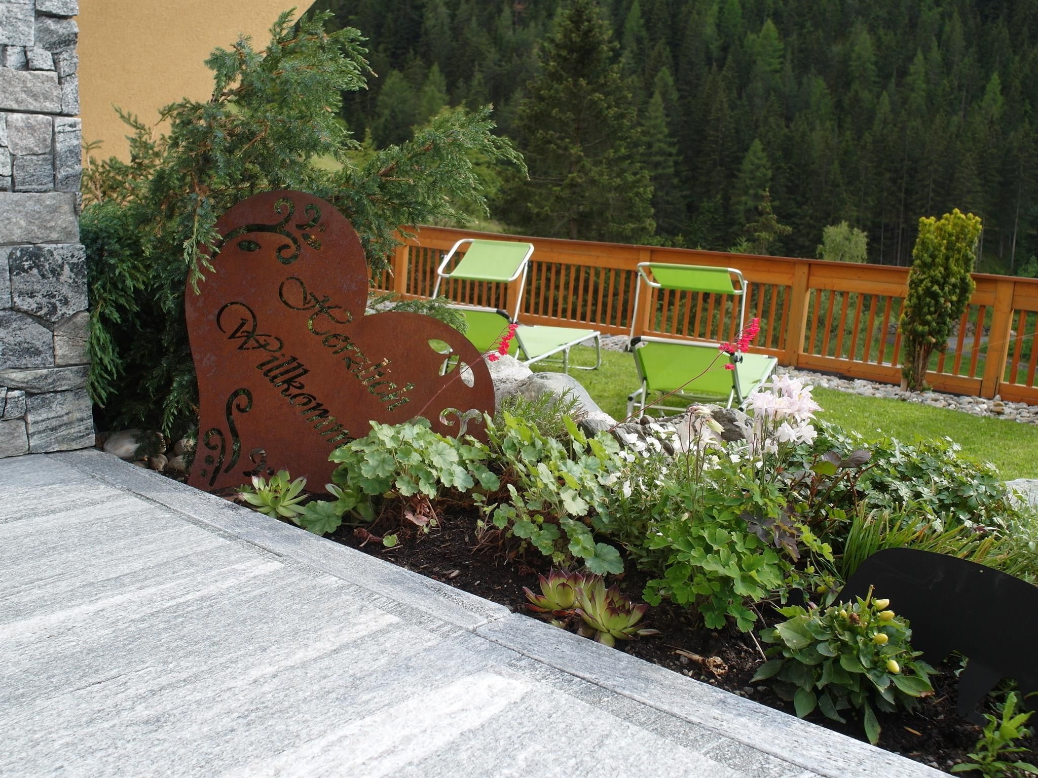 An inviting garden with a heart-shaped welcome sign. In the background, there are comfortable loungers and numerous plants.