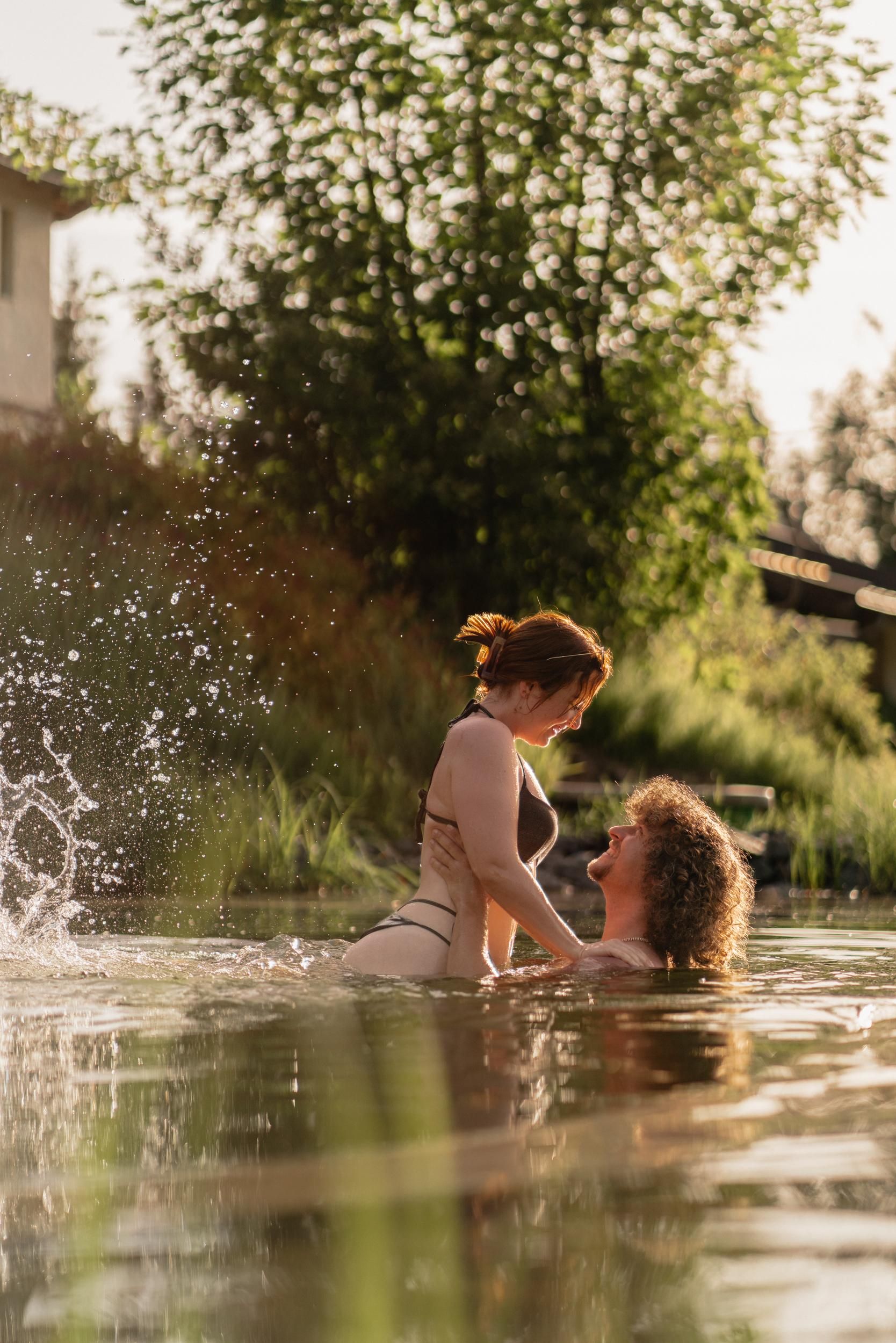 A couple stands in the water and looks at each other. It is a sunny setting with green plants in the background.