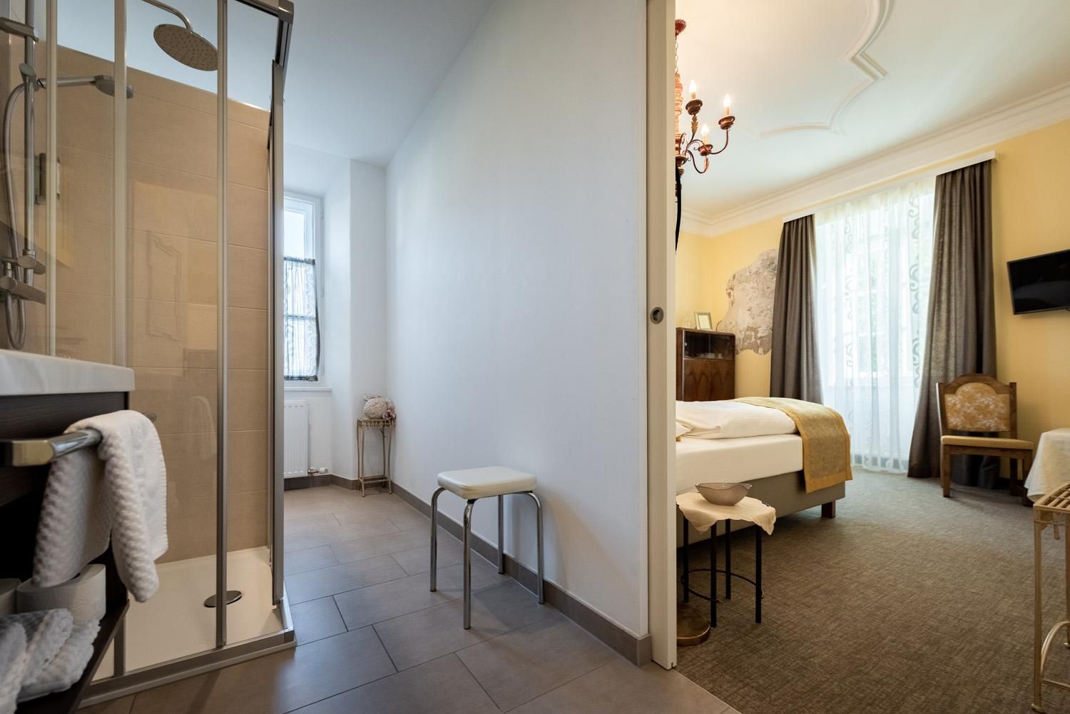 A modern bathroom with a shower and towels. In the background, a cozy bedroom with windows and a bright atmosphere can be seen.