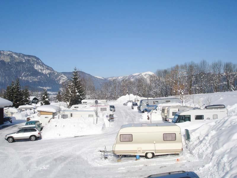 A campsite in winter with caravans, surrounded by snow and mountains. The sky is clear and blue.