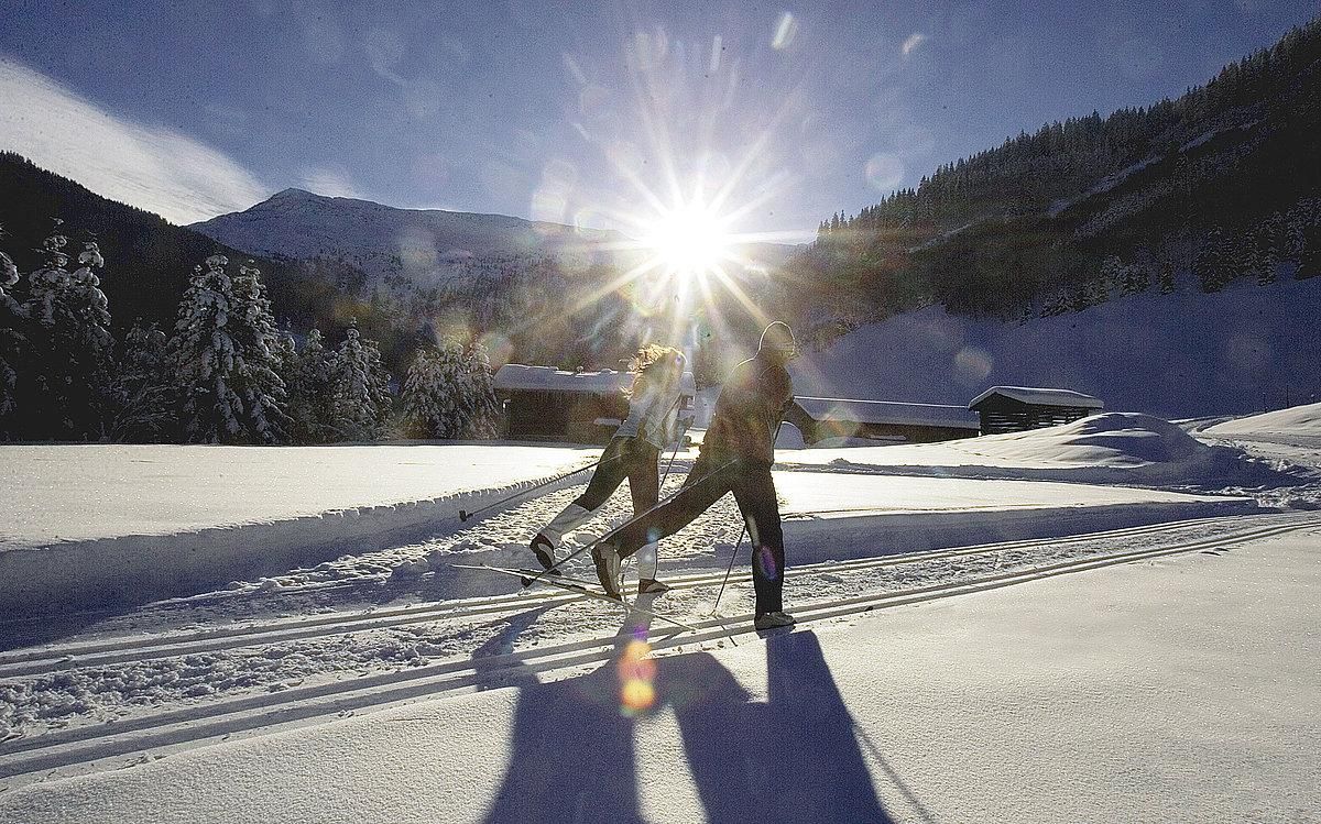 Two people are cross-country skiing through a snowy landscape. The sun shines brightly over the mountains, creating a beautiful winter atmosphere.