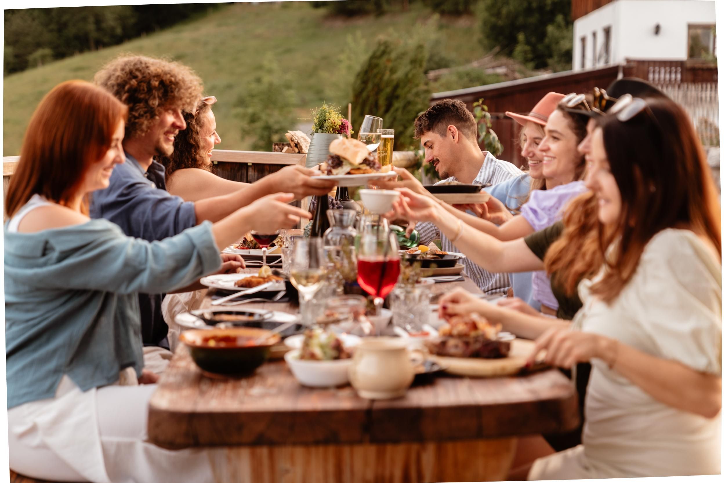A friendly group of people is sitting at a wooden table, enjoying a festive meal outdoors. They are sharing dishes and toasting with drinks while the sun is shining.
