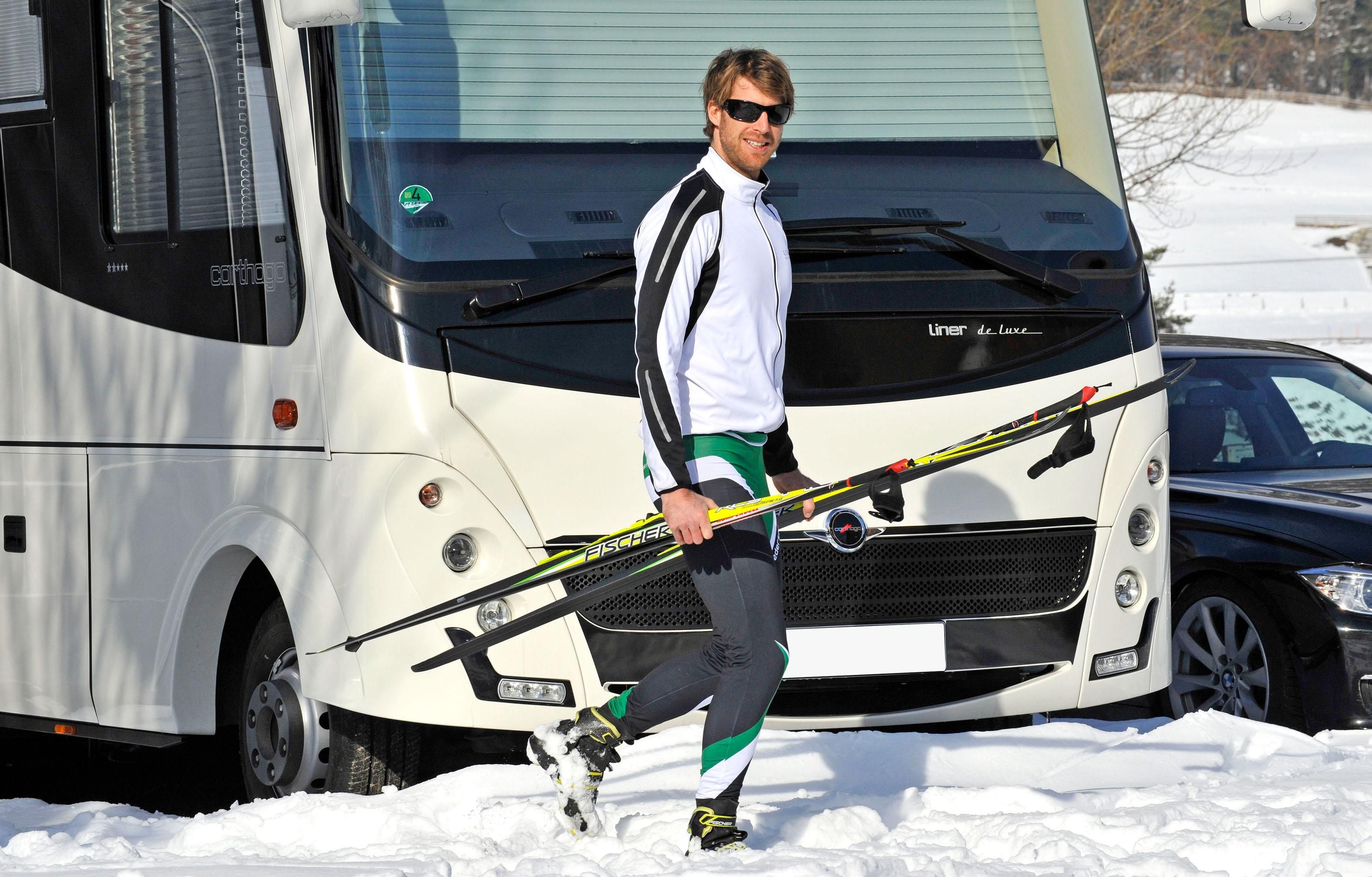 A skier is carrying his equipment and walking on snow to a motorhome. In the background, other vehicles can be seen.