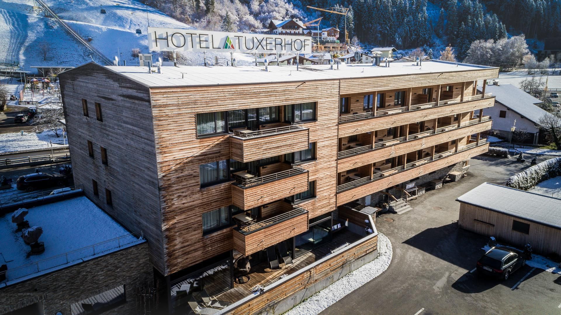 A modern hotel building with a wooden facade and several balconies. The surroundings are snow-covered, and mountains are visible in the background.