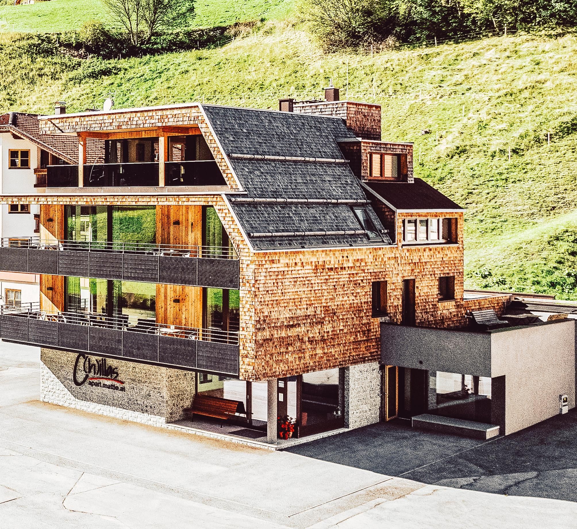 A modern building with a wooden and stone facade set in a green landscape. It features large windows and balconies that create a bright and inviting atmosphere.