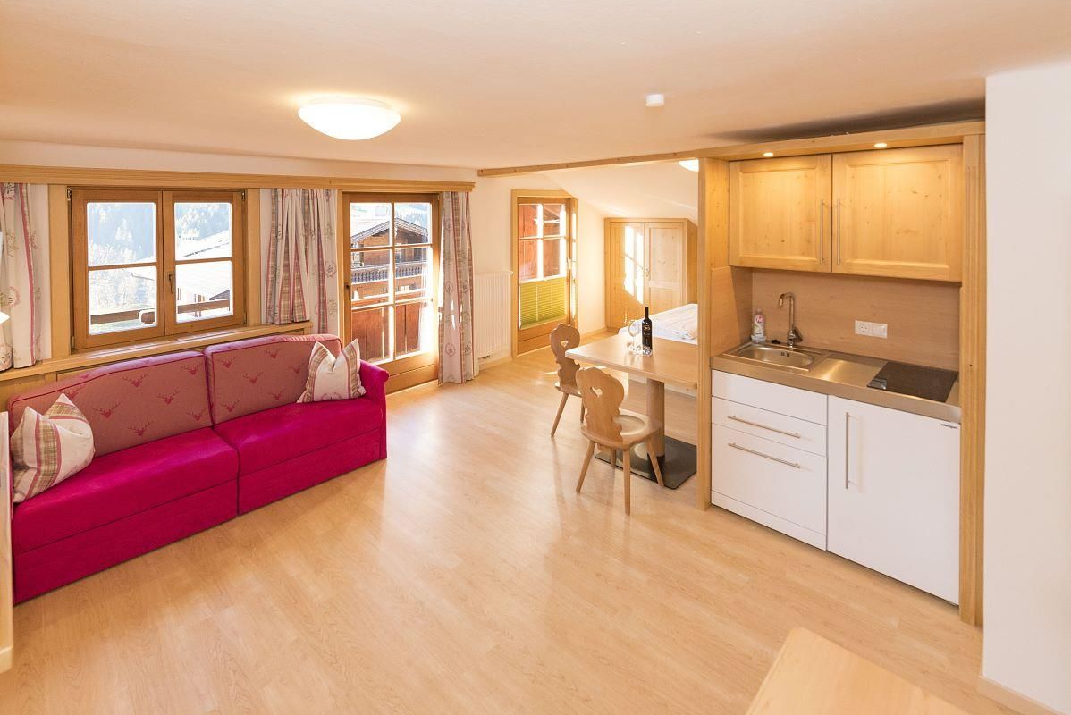 A modern, bright apartment with a red sofa and a kitchenette. Large windows provide plenty of daylight and a cozy feel.