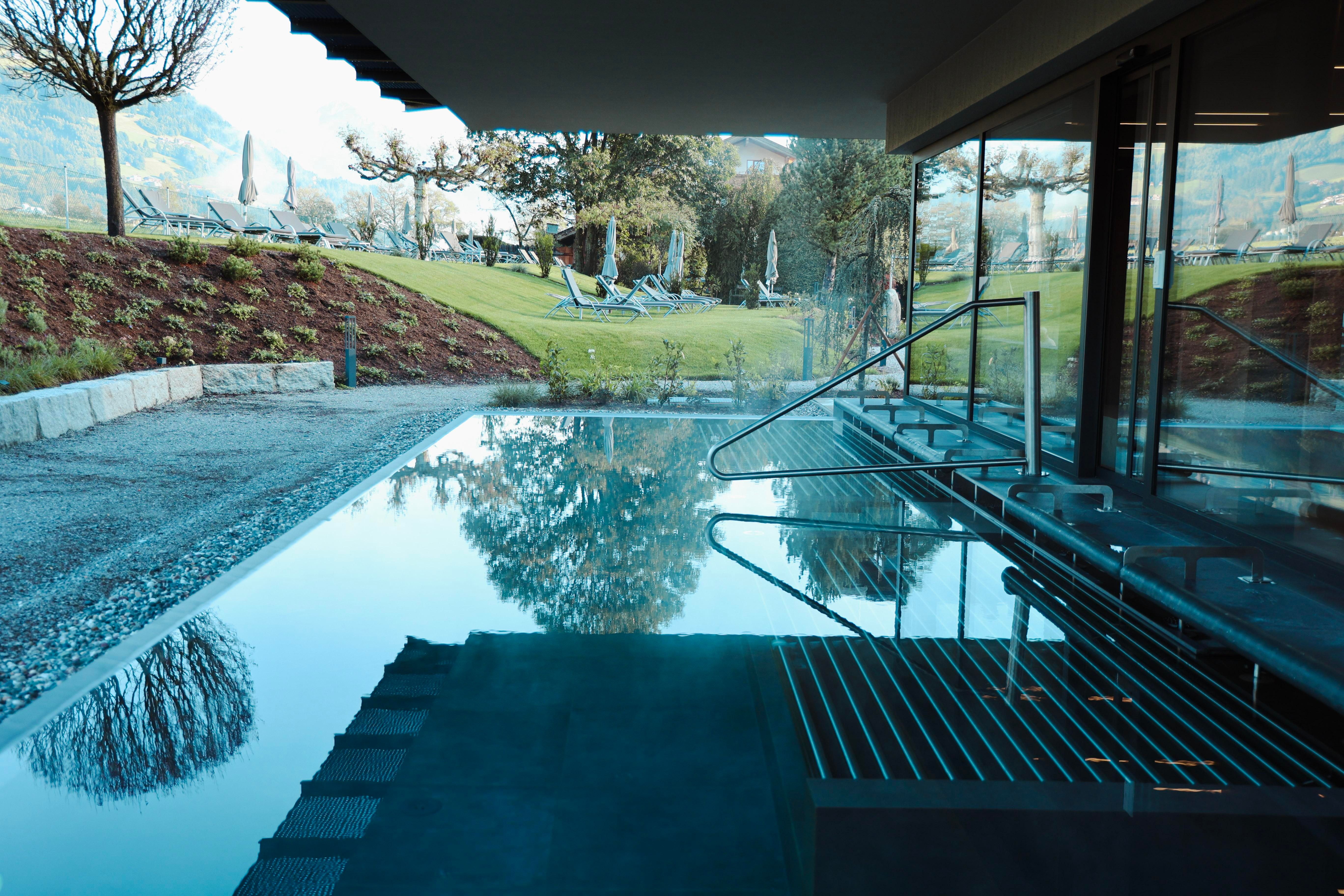 An elegant spa with a clear water pool and a view of a green meadow. In the background, there are relaxing loungers and trees visible.