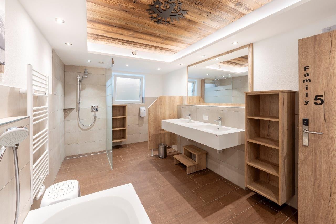 A modern bathroom with a wooden ceiling and large mirrors. It features a shower, a double sink, and shelves for storage.