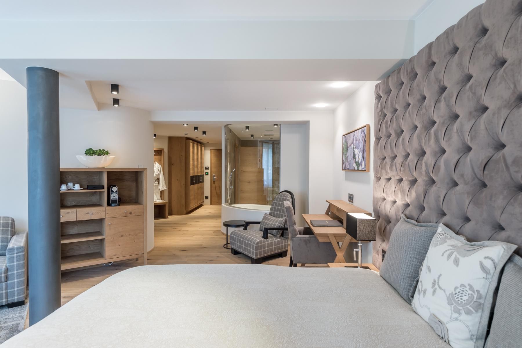 A modern bedroom with a large, upholstered headboard and a cozy chair. The room is bright and well-designed, featuring wood and light-toned accents.