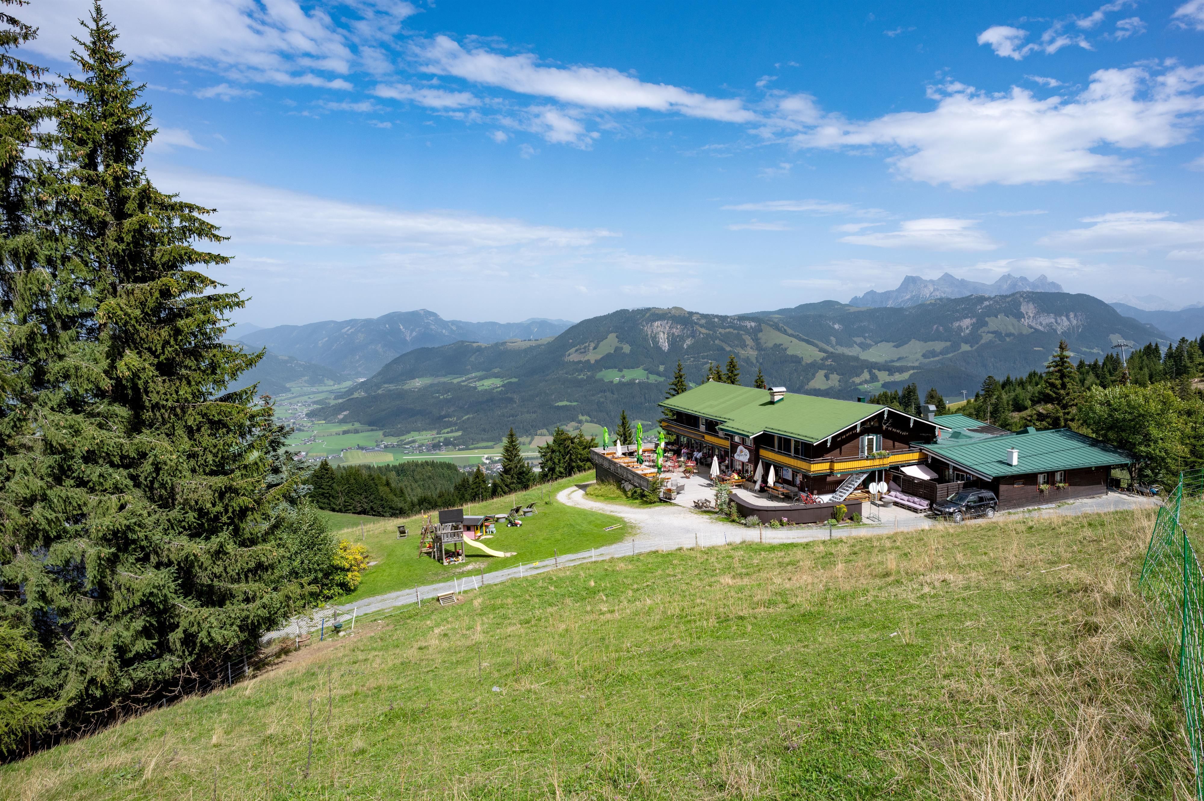 A picturesque mountain landscape with a cozy restaurant in the foreground. In the background, green hills and a clear blue sky can be seen.