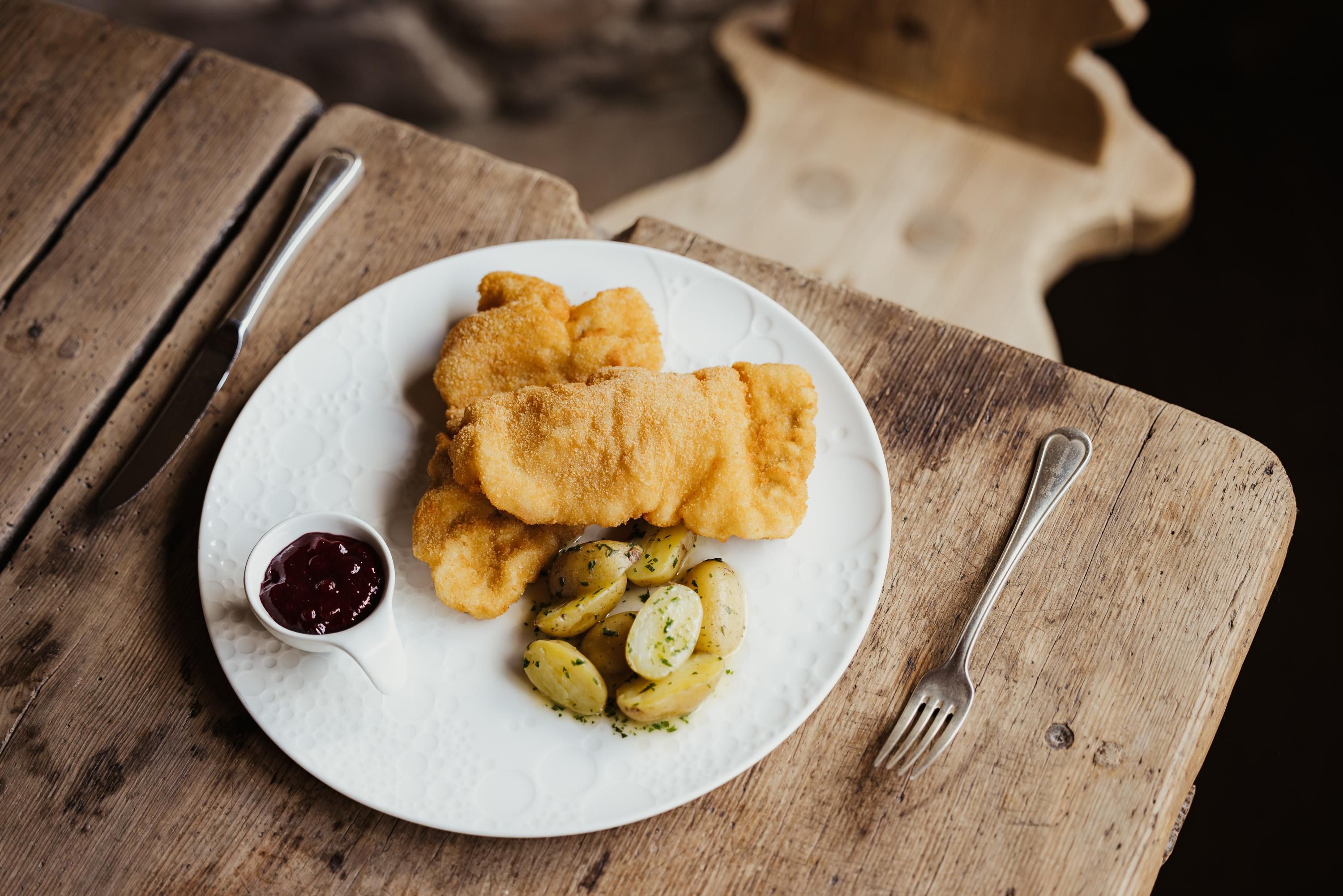 A plate with golden-brown fish, served with potatoes and a sauce. The plate is on a rustic table.