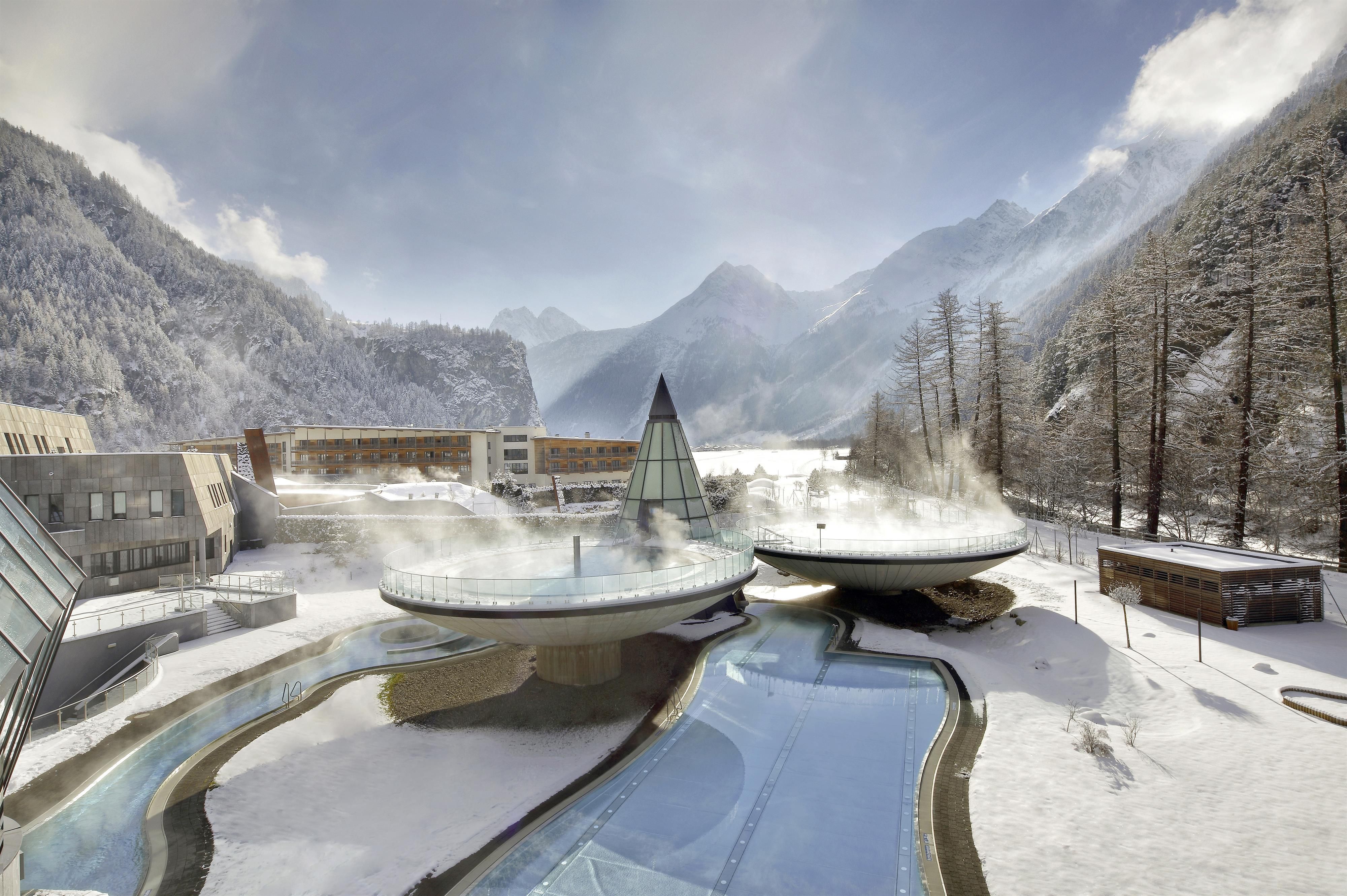 An impressive winter landscape with snow-covered mountains and a modern spa complex. Columns of steam rise from the thermal pools, surrounded by a tranquil natural backdrop.