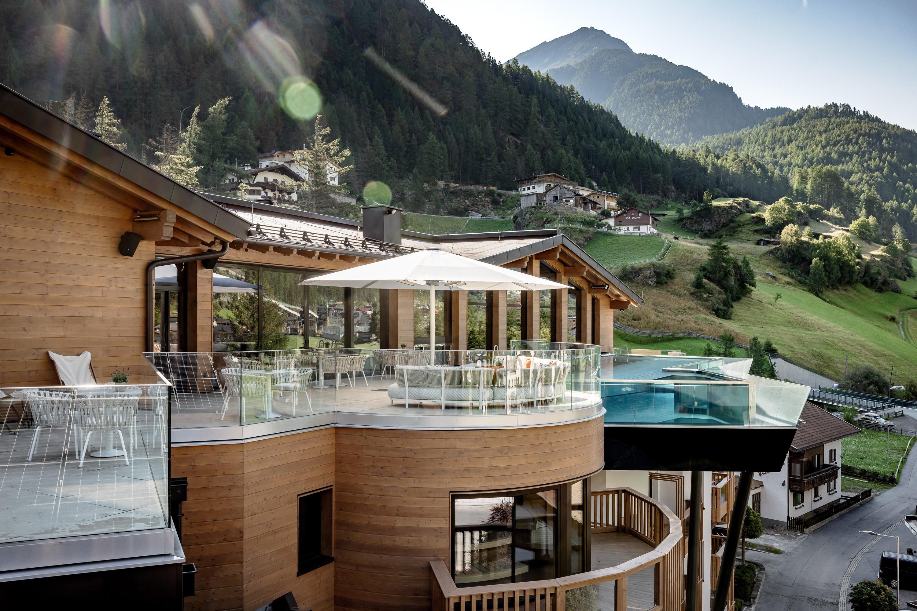 A modern chalet with a wooden facade and balcony, surrounded by green mountains. In the foreground, there is a pool and a terrace with a sun umbrella.