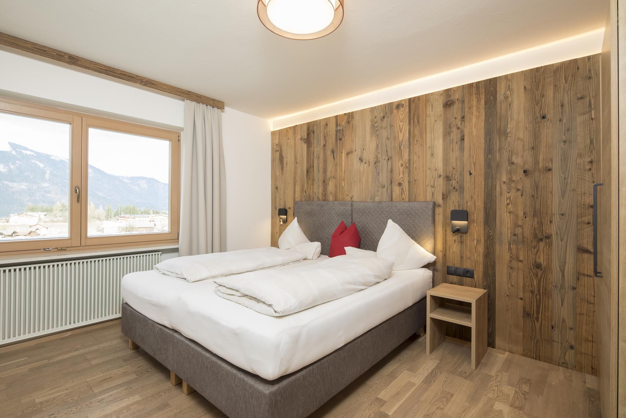 A modern bedroom with a double bed and wooden paneling on the wall. Large windows offer a view of the mountains.
