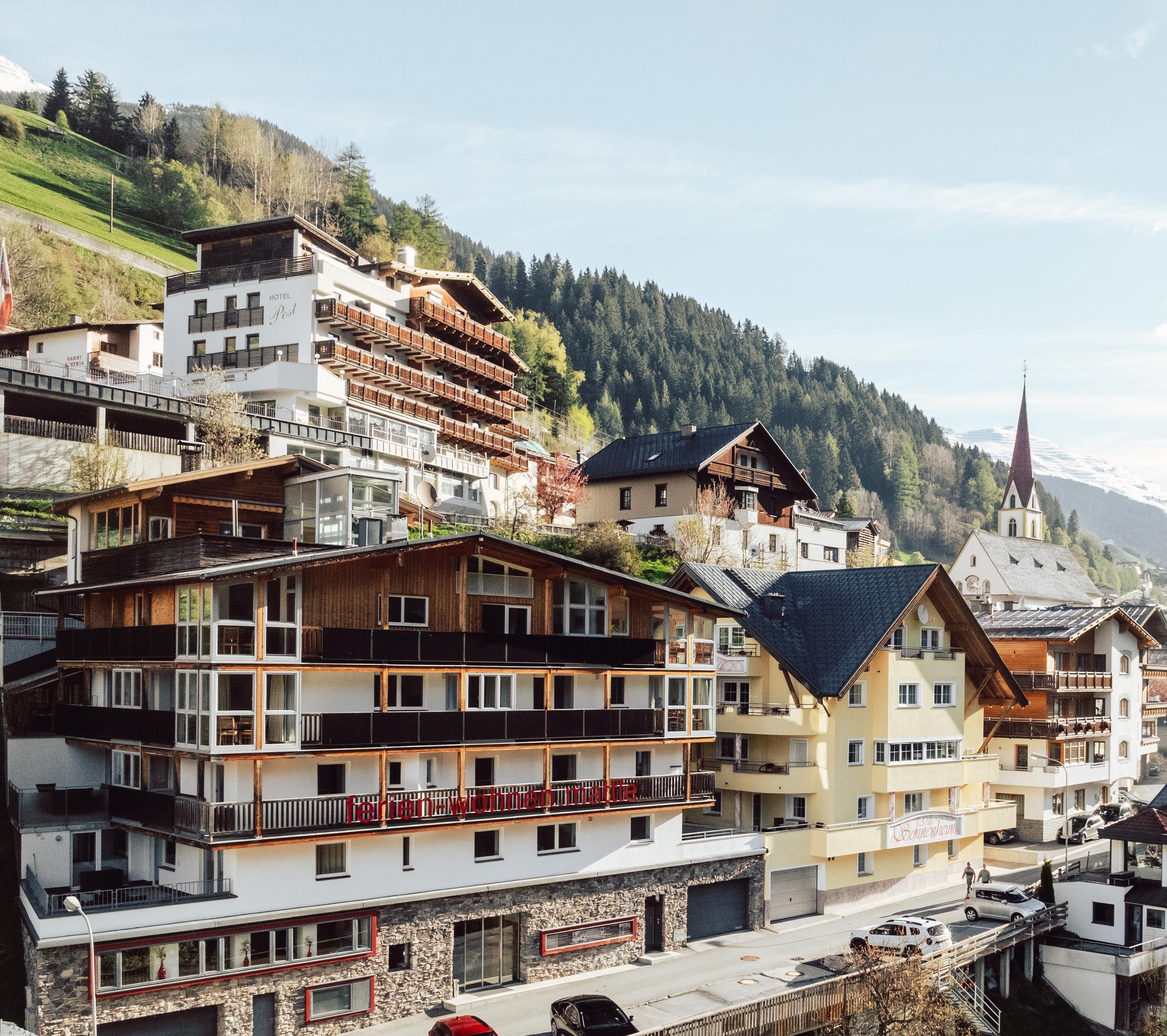 A picturesque mountain landscape with several modern chalets and traditional buildings. The houses are situated on a slope, surrounded by forests and mountains.