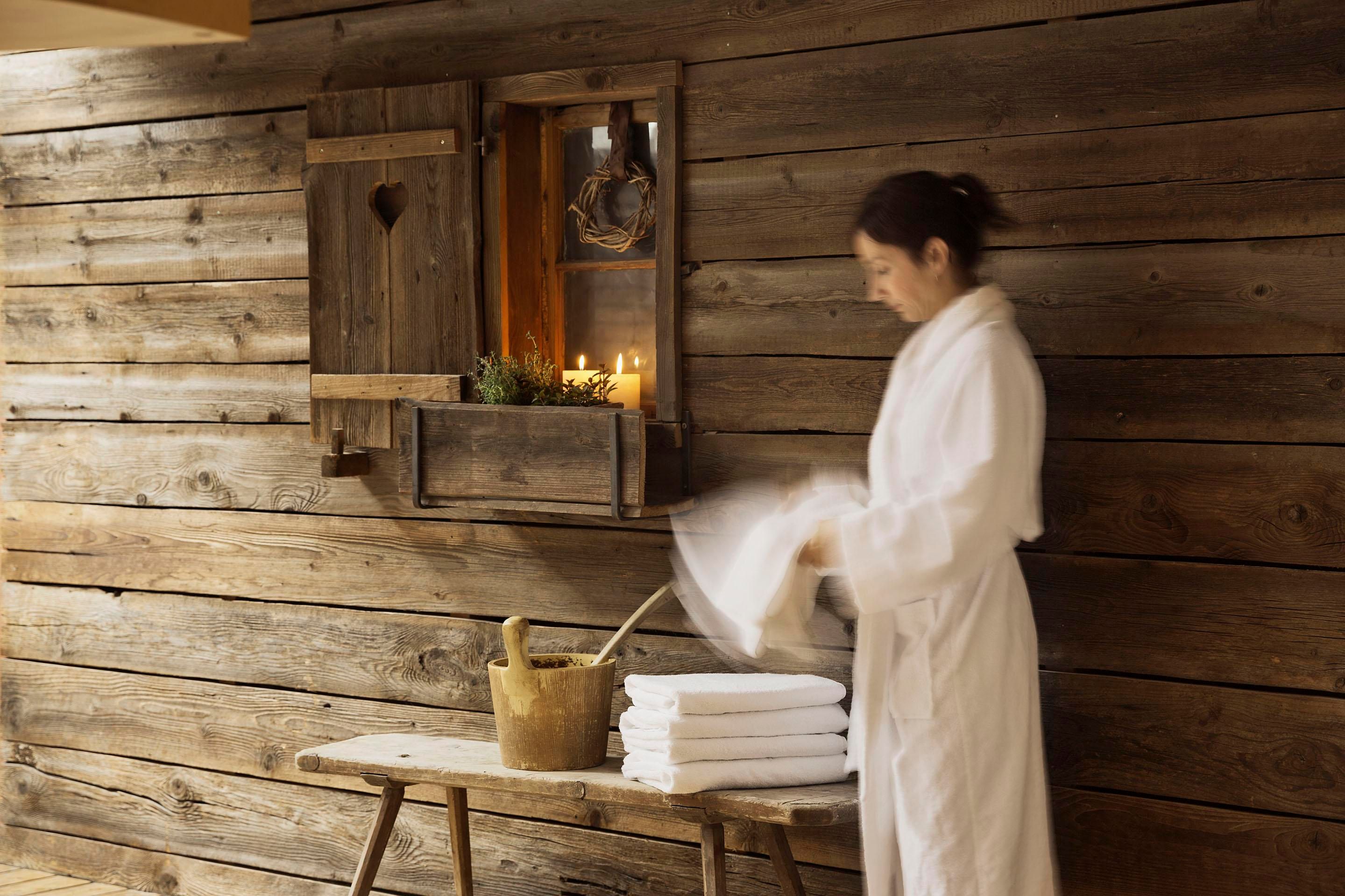 A woman in a bathrobe is preparing in a wellness area. The room has a rustic wooden wall and candlelight creates a relaxed atmosphere.