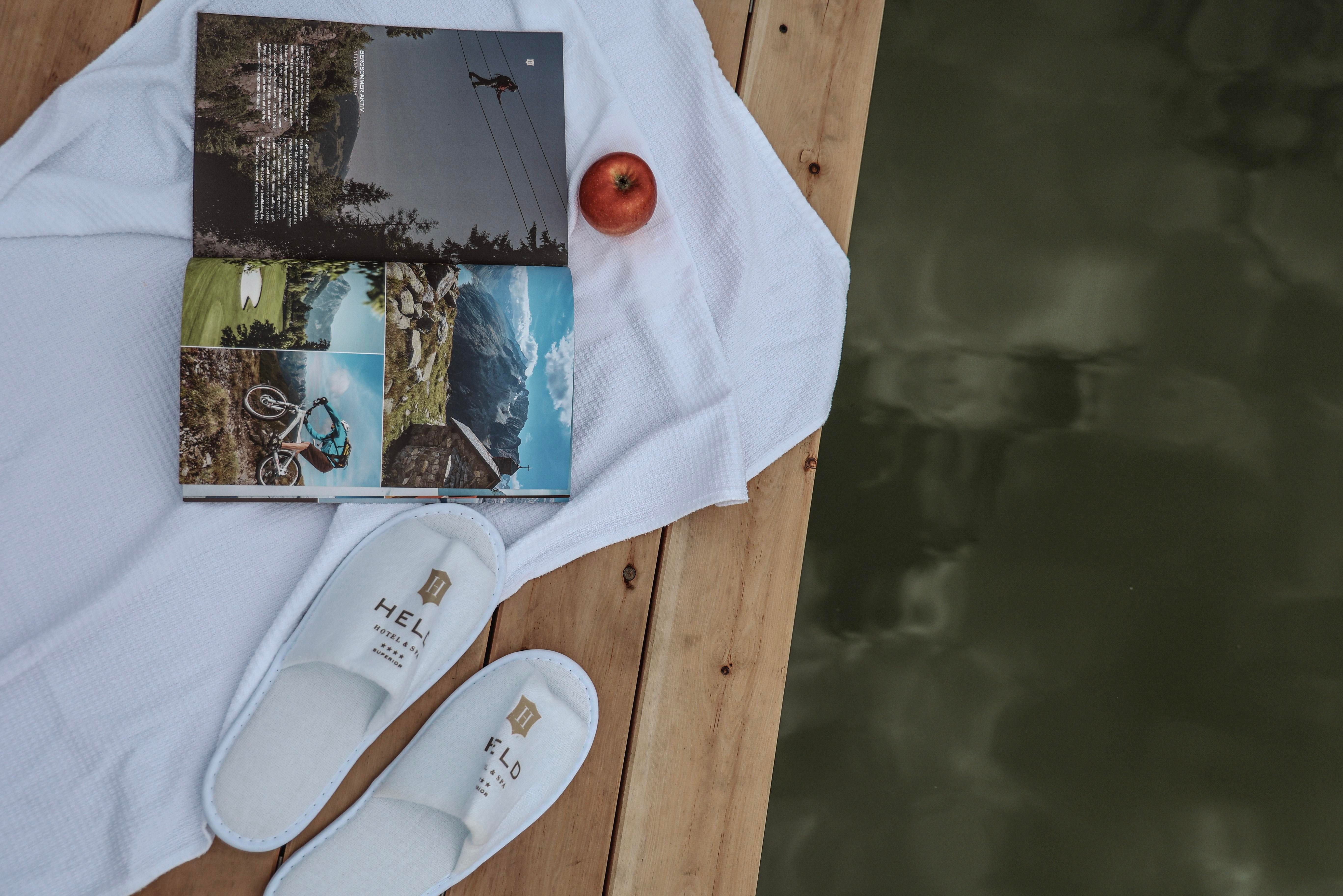 A towel lies on a wooden deck next to a book, an apple, and white house slippers. In the background, the water is reflecting.