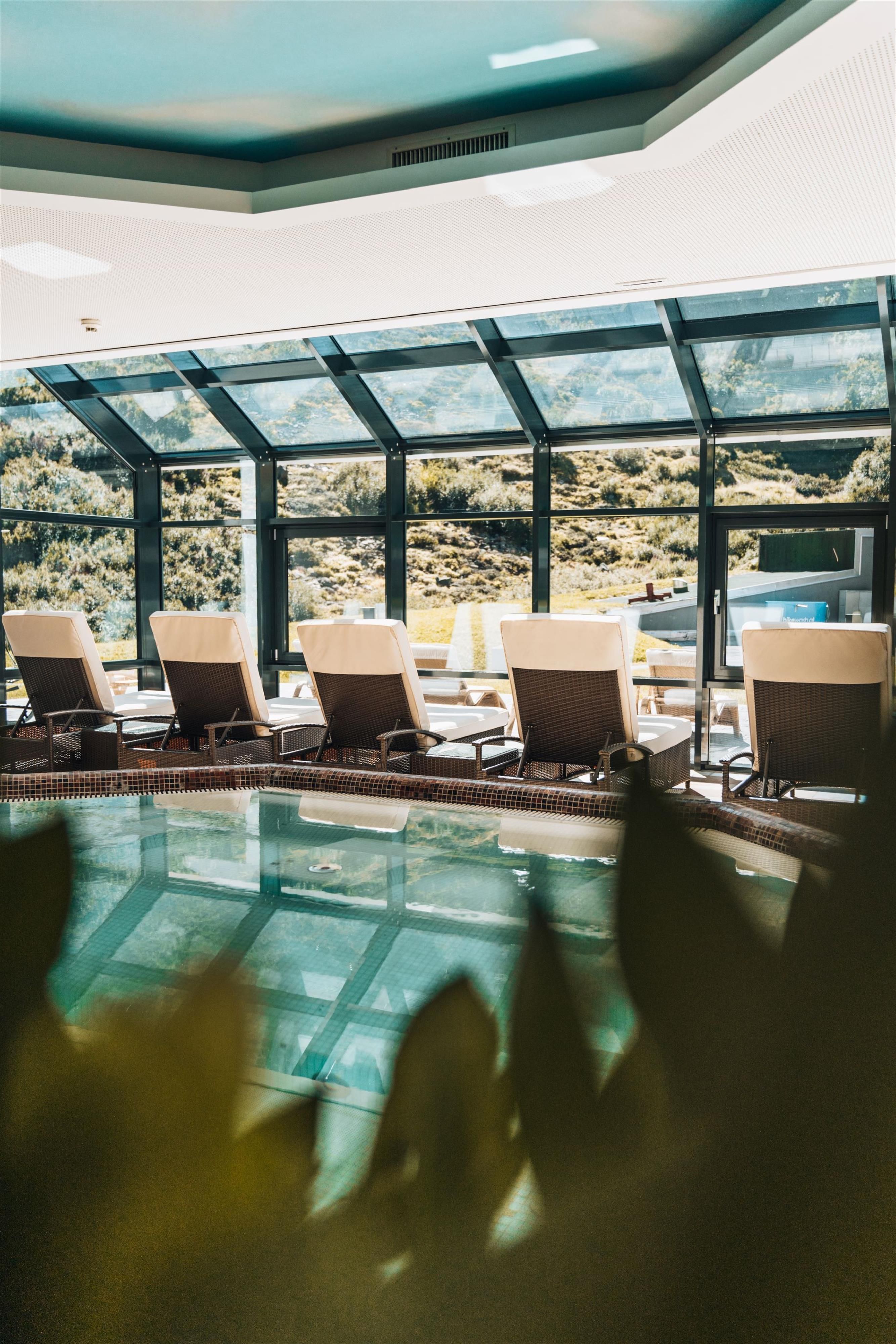 An elegant indoor pool area with loungers and large windows that offer a view of the outside. The ambiance is bright and modern.